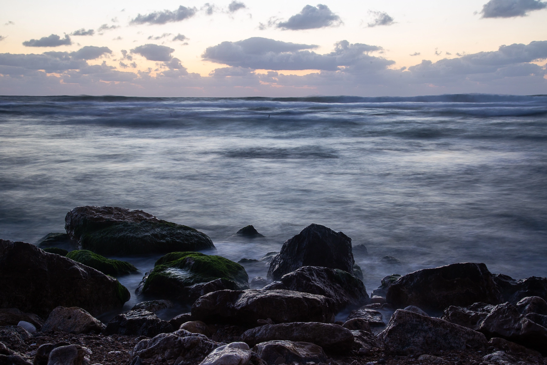 Waves crashing against moss-covered rocks at the shore, as the sun sets behind a cloudy sky, casting a serene glow over the ocean. Sidney Ali Beach, Israel