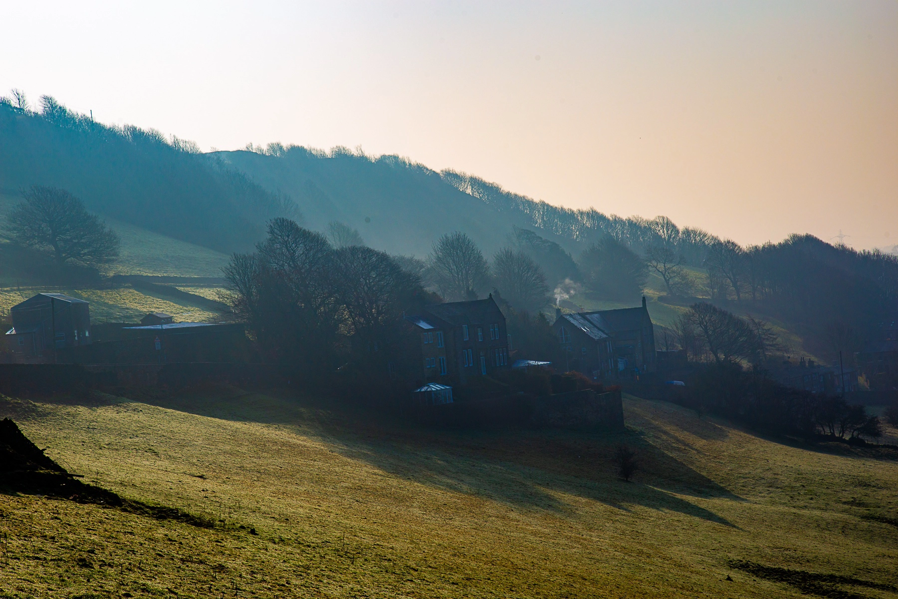 A misty morning view of rolling hills with stone houses nestled in a valley, soft sunlight casting long shadows over the green fields. Halifax, Great Britain