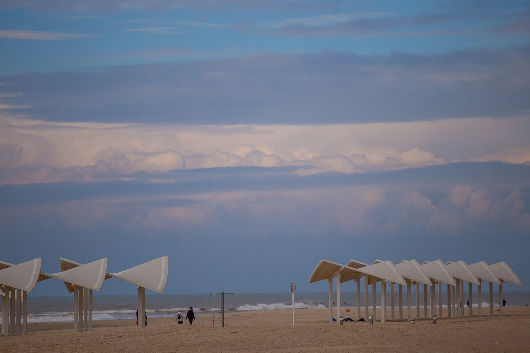 Modern white canopy structures on a quiet beach with soft waves, a cloudy sky, and a few people strolling by. Hertzliya, Israel