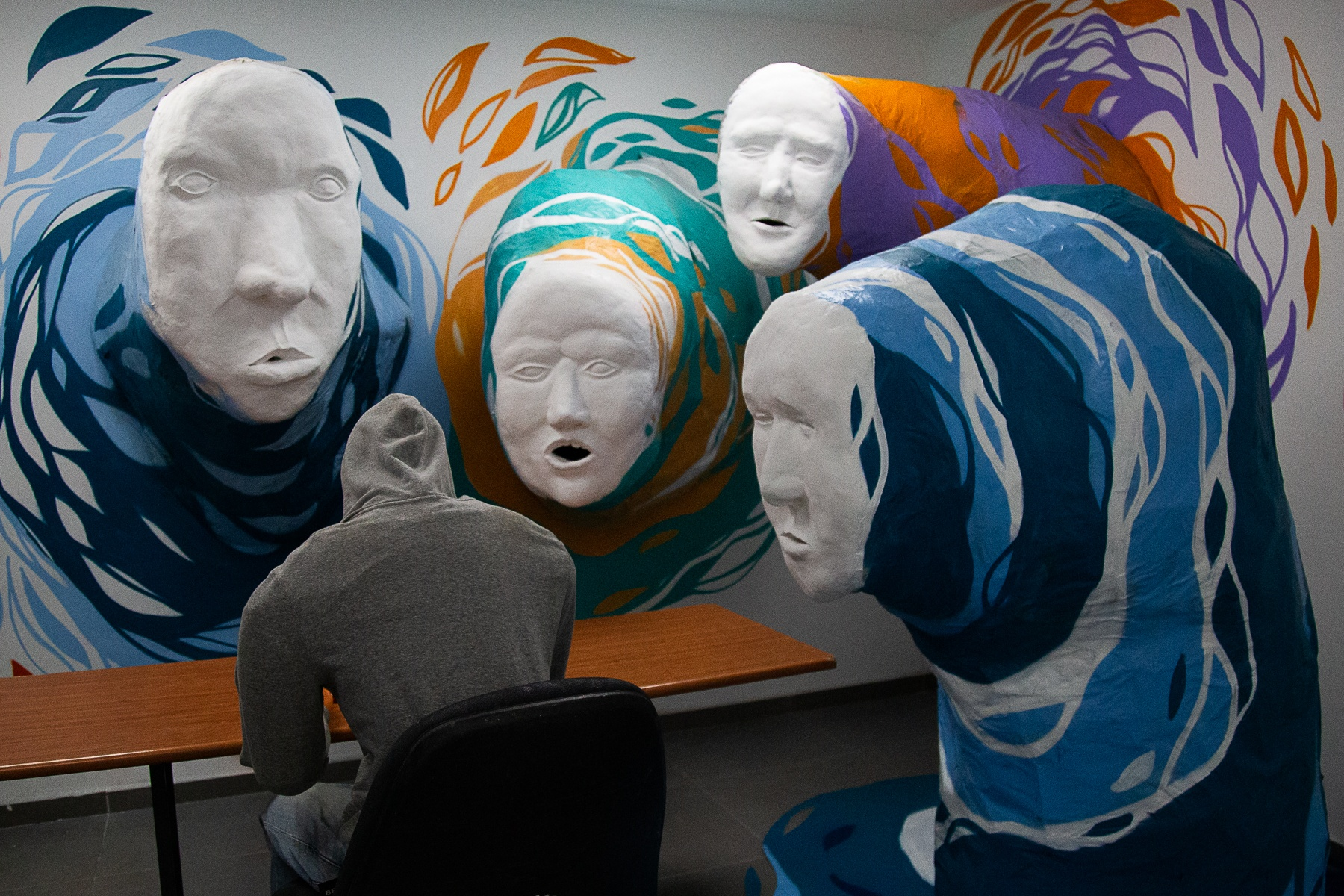 A hooded figure sits at a table, surrounded by large, expressive faces in vibrant swirling patterns.