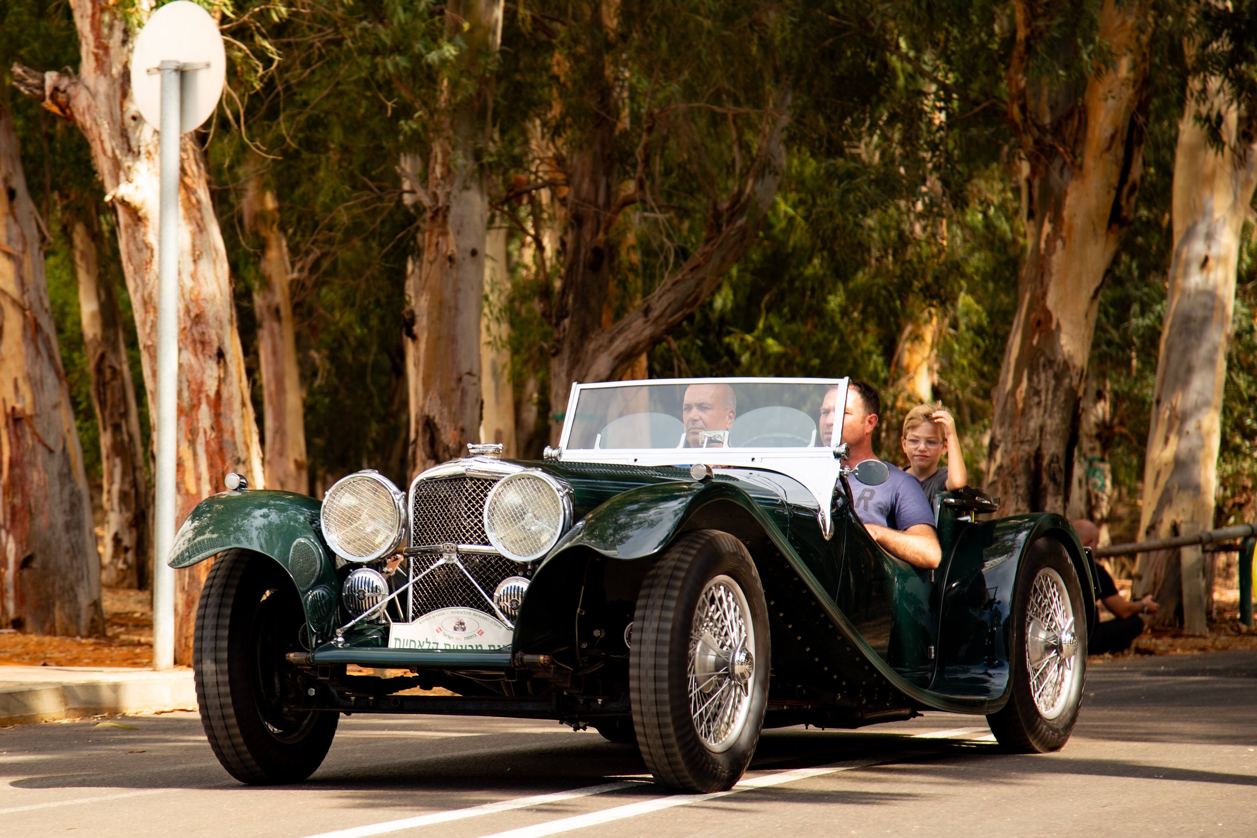 A classic green vintage car with two passengers driving through a tree-lined road, part of a retro car event. Retro Car Club Leumi, Tel Aviv, Israel