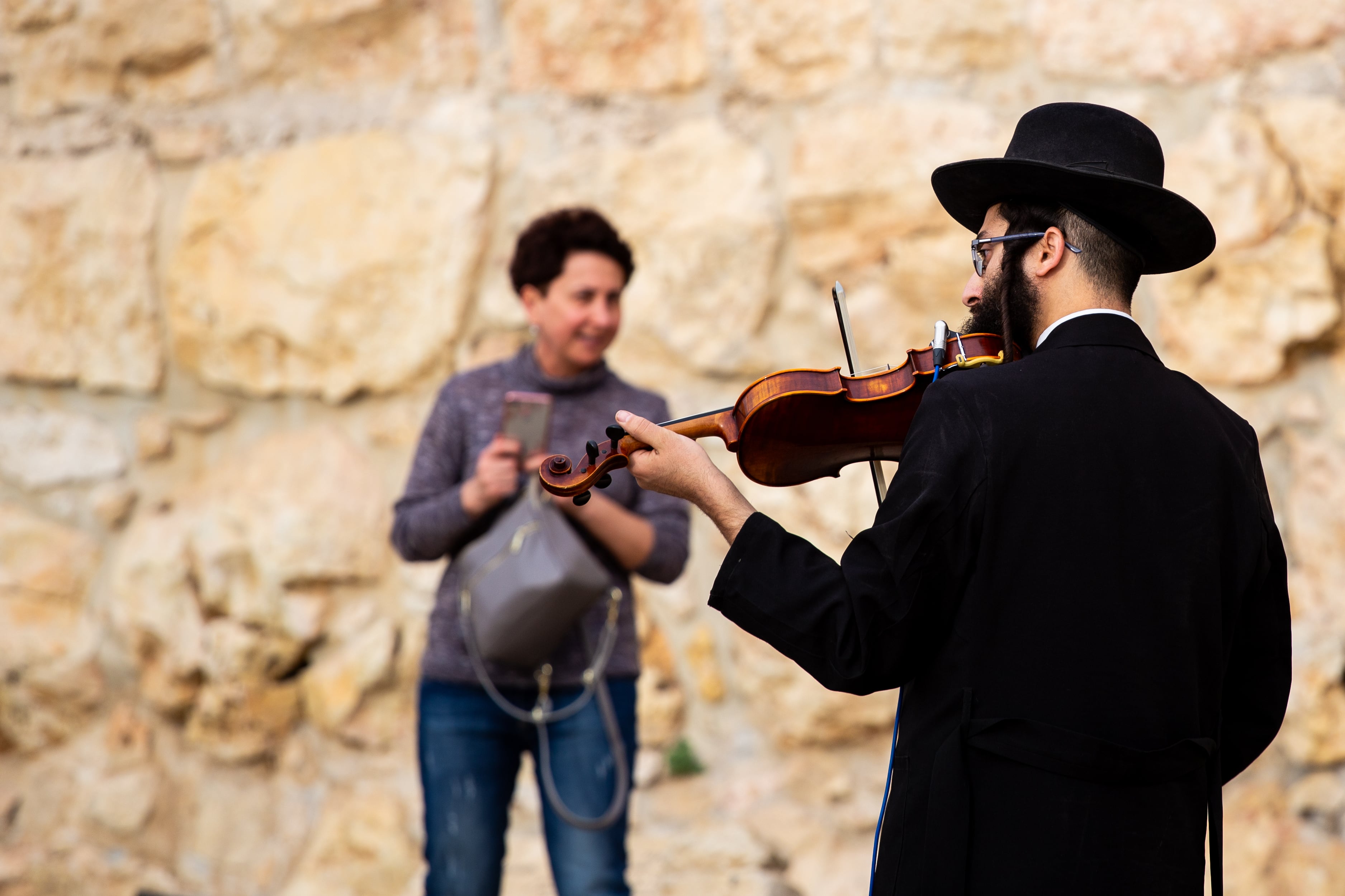 A violinist plays in Jerusalem’s Old City, captivating a passerby near a stone wall, adding to the timeless feel. Old City, Jerusalem, Israel