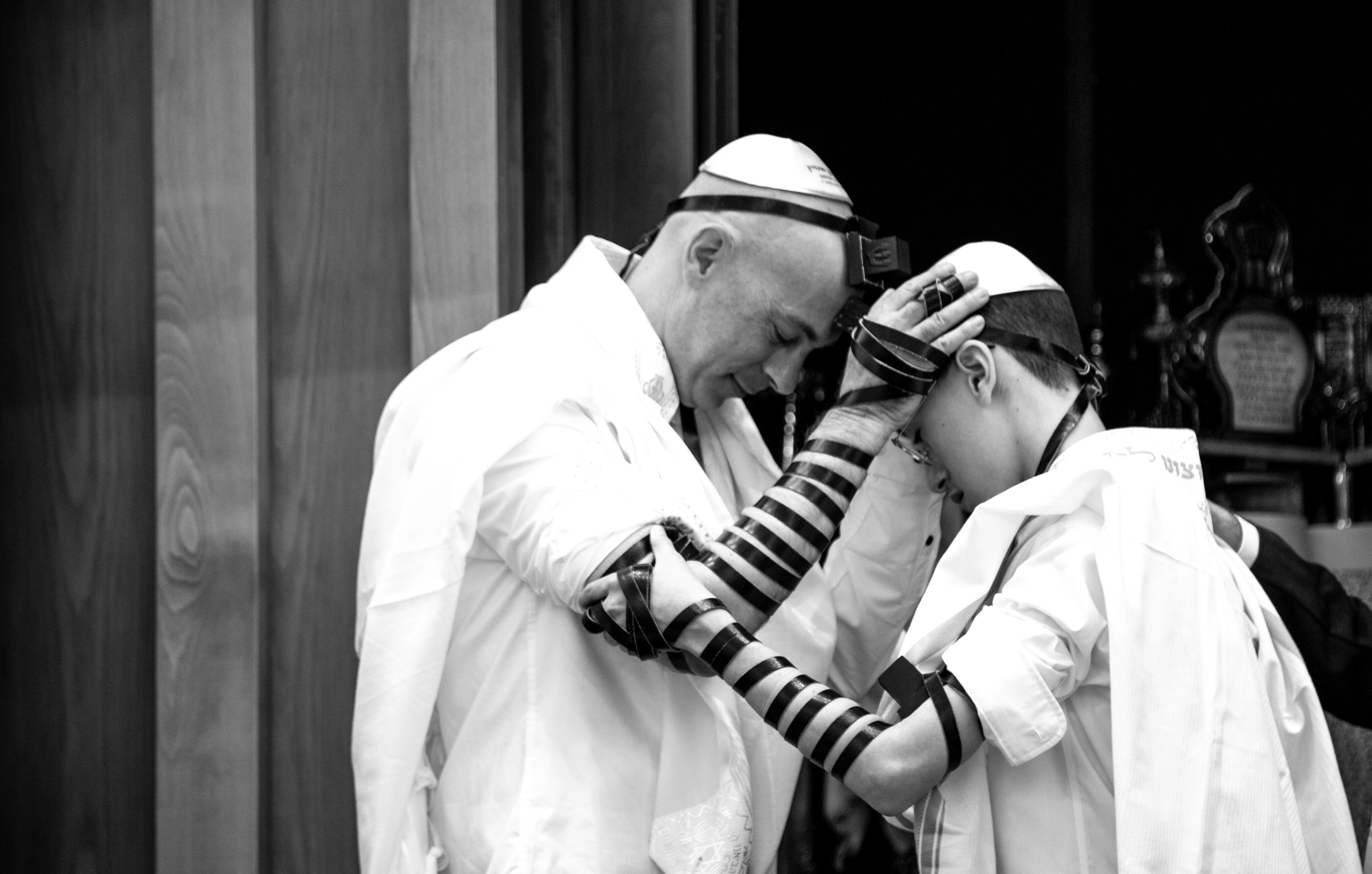 A black-and-white photo of a father and son in prayer, wearing tefillin, sharing an intimate connection. Raanana, Israel