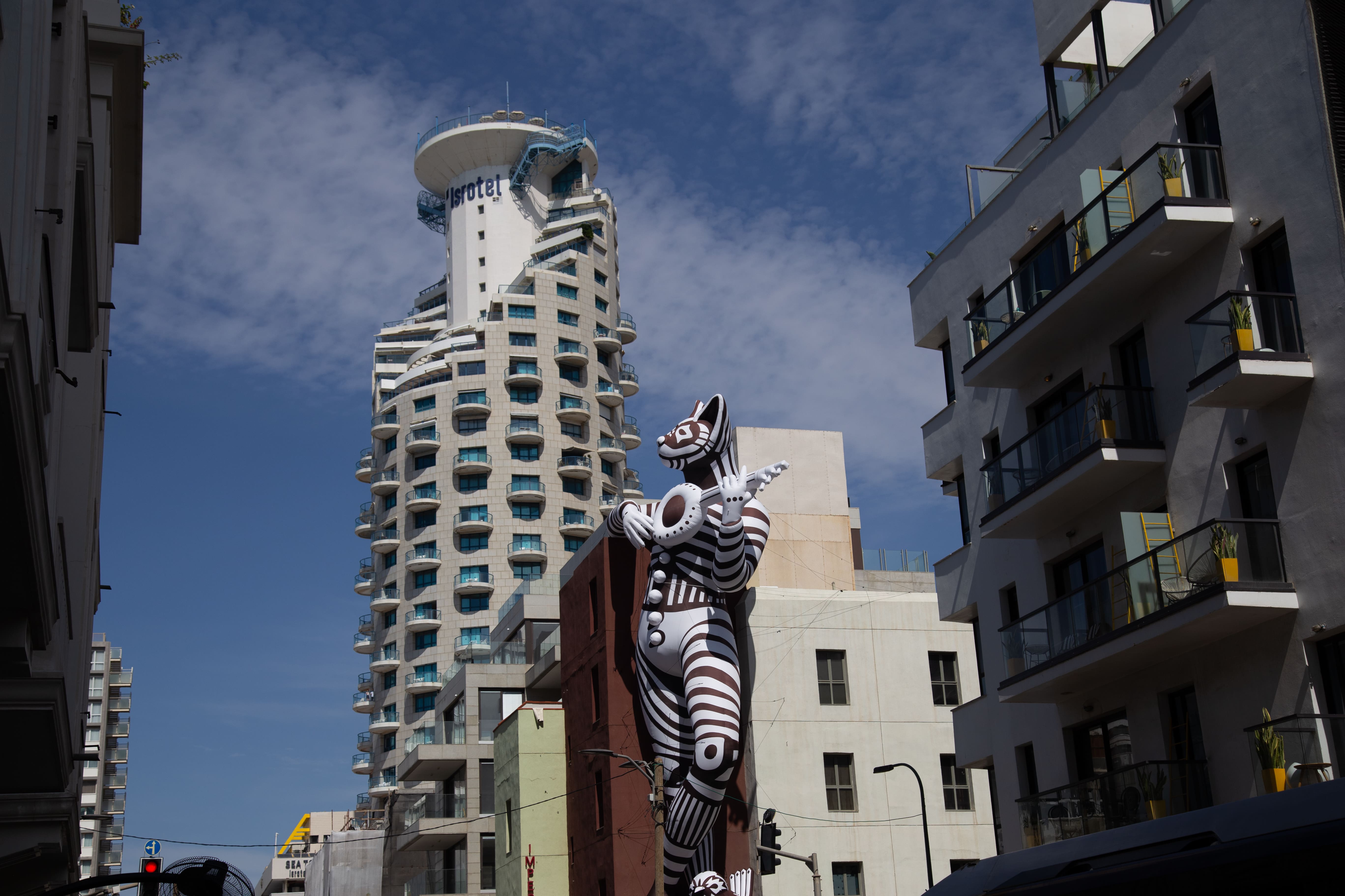 A towering, striped cat figure plays a wind instrument in a surreal urban setting, blending whimsy with sleek, otherworldly elements. Tel Aviv Imperial Hotel