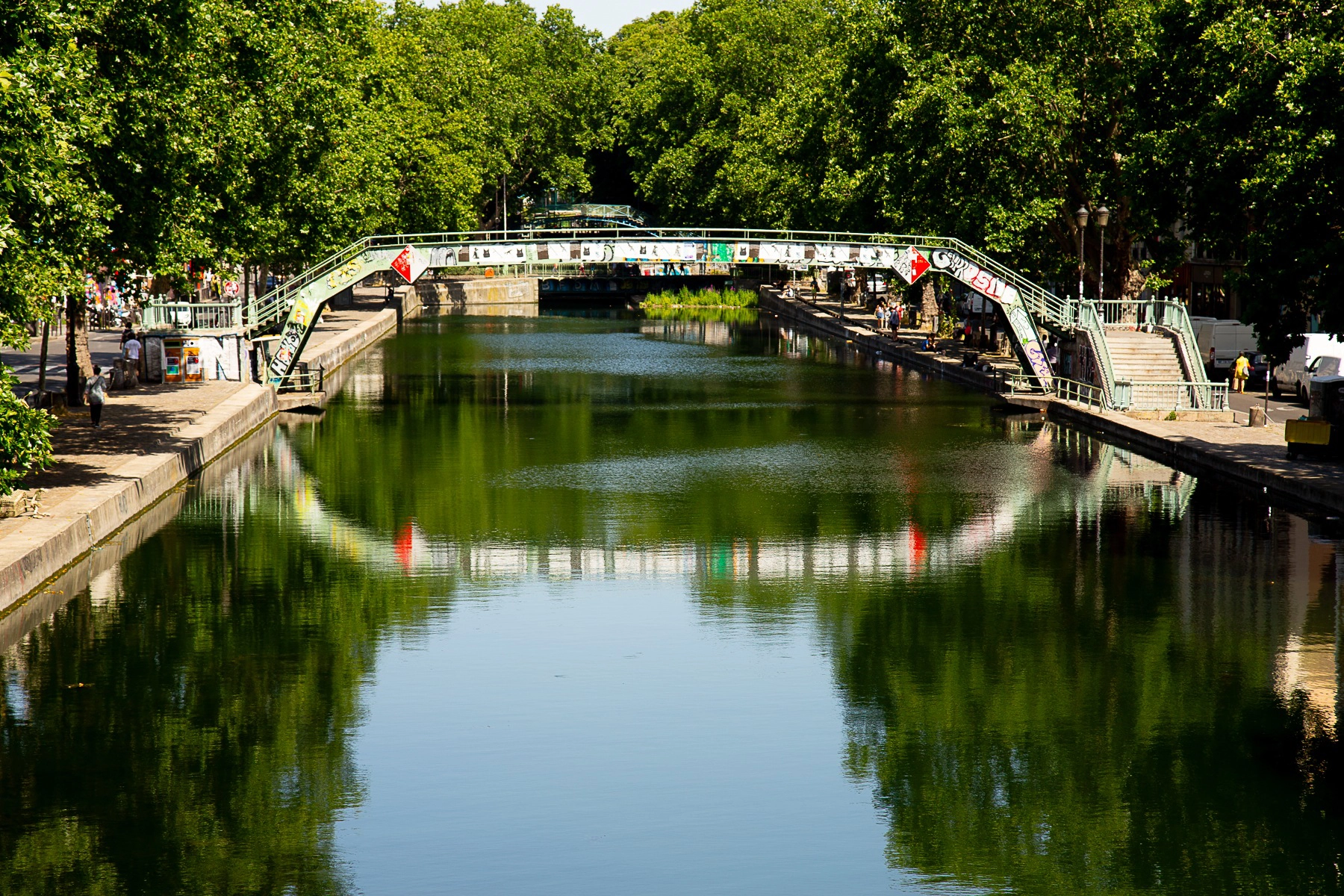 A view of the green waters of Canal St-Martin in Paris, framed by lush trees, with a quaint pedestrian bridge in the distance. Paris, France