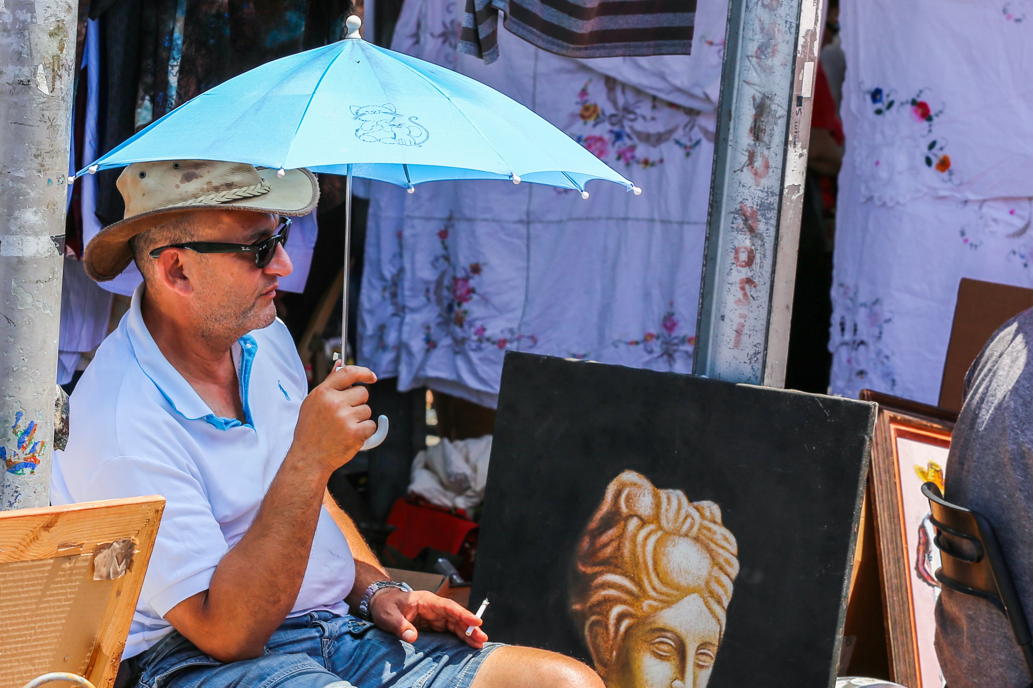 A vendor relaxes under a blue umbrella at Jaffa Flea Market, next to a classical statue painting. Jaffa Flea Market, Tel Aviv, Israel