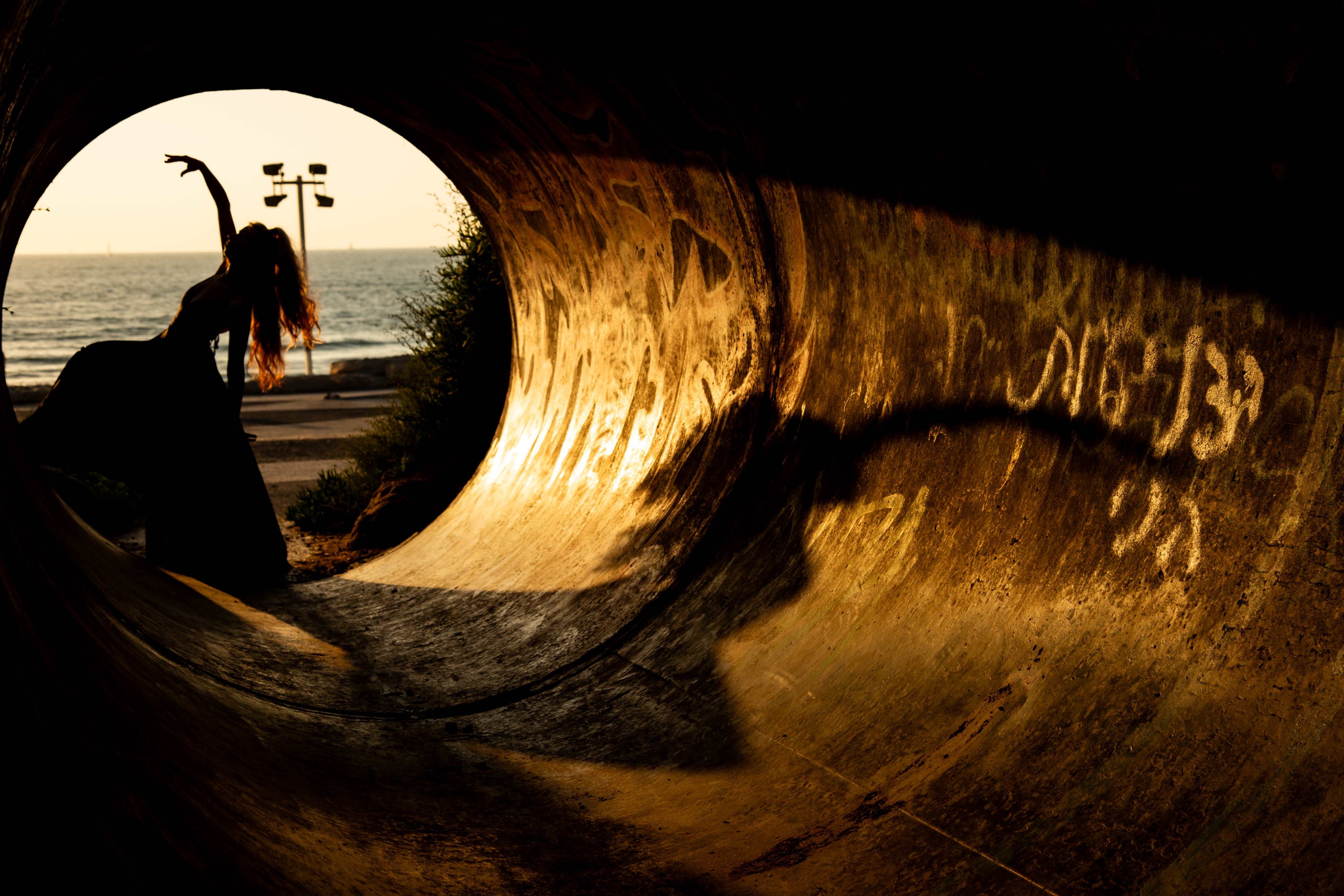 A silhouetted figure striking a graceful pose at sunset, framed inside a circular tunnel by the sea. Tel Aviv, Israel