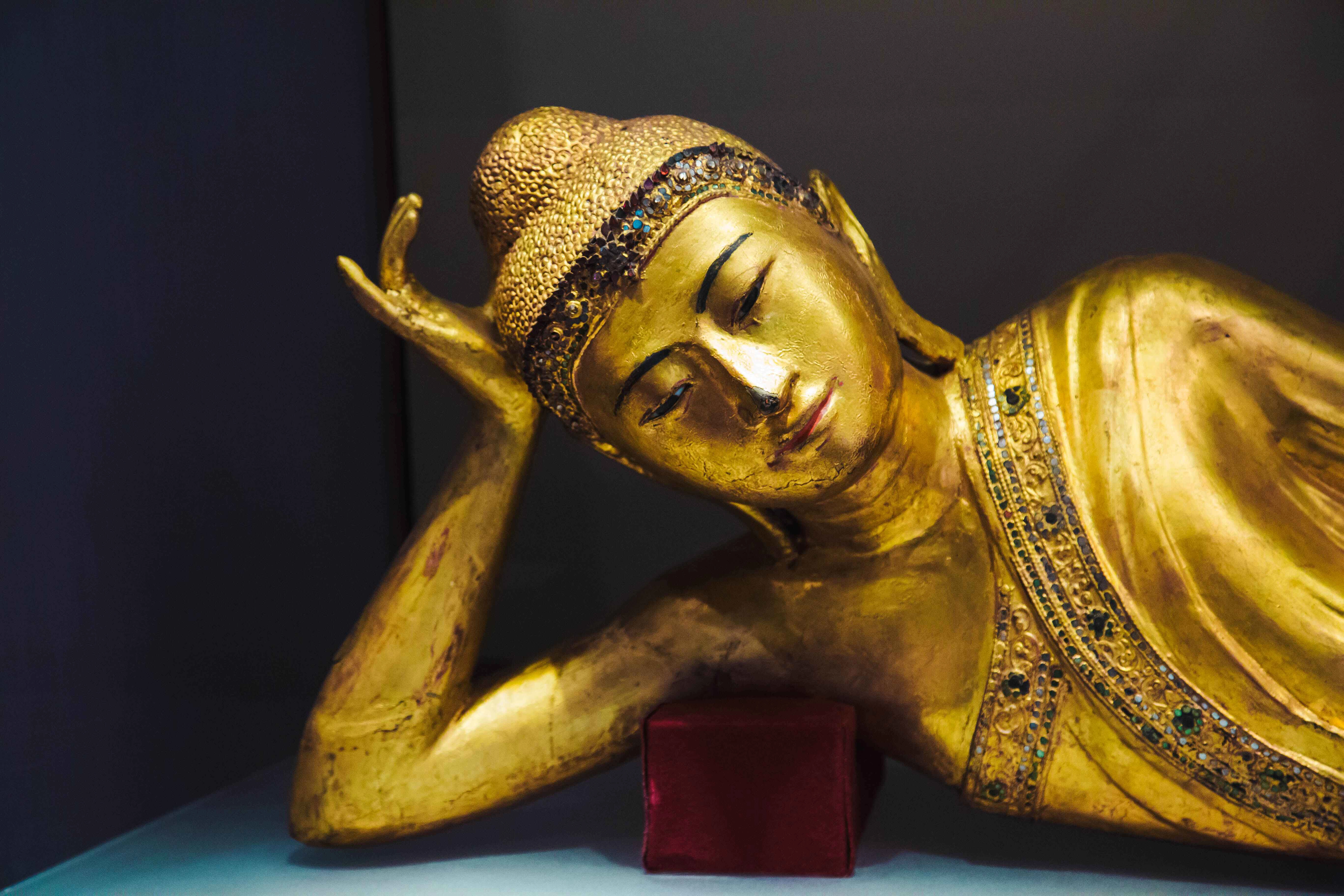 A golden reclining Buddha statue, adorned with intricate details and jewels, resting on a red cushion. Turin, Italy