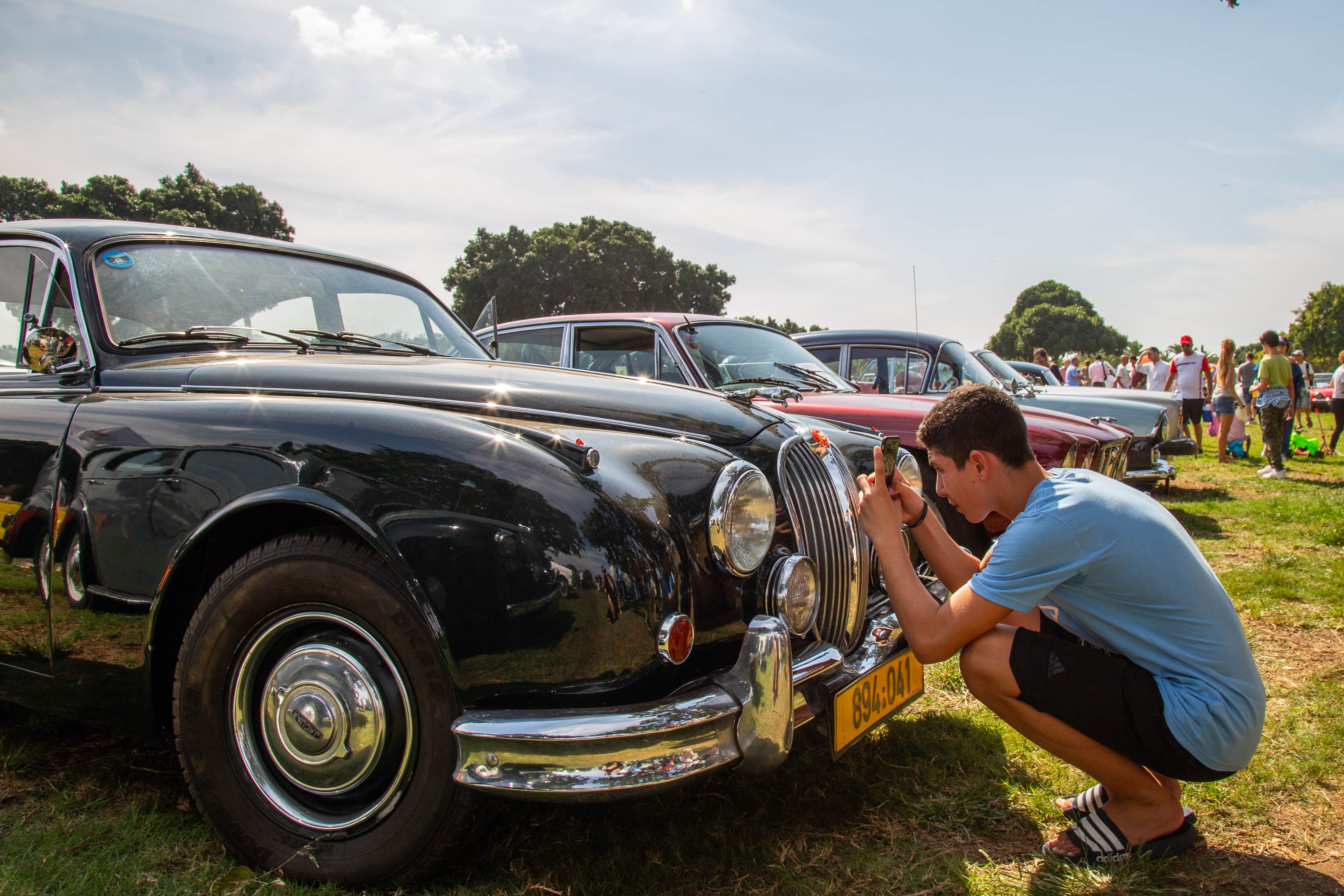 A young photographer crouches to shoot a vintage car at a Retro Car event, celebrating automotive history. Retro Car Club, Leumi Park, Israel