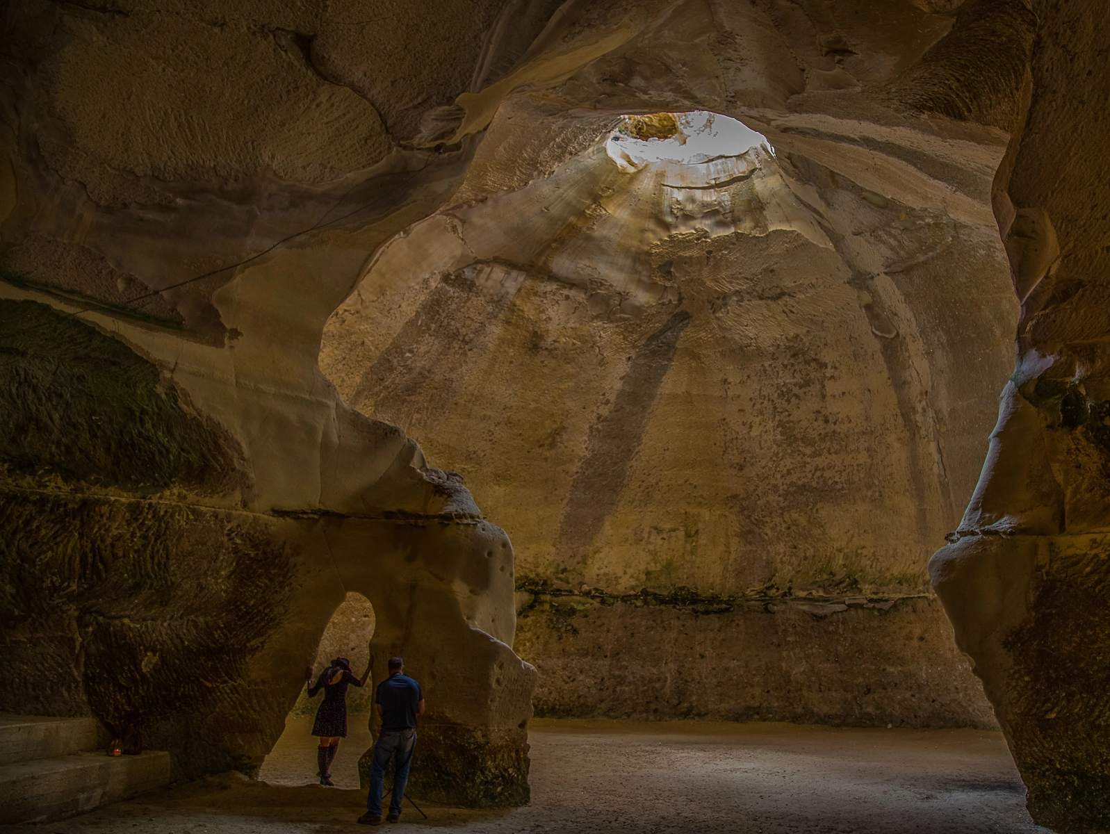 Two people standing in a large cave with sunlight beaming through a circular opening above, casting a mystical light on the stone walls. Beit Guvrin, Israel