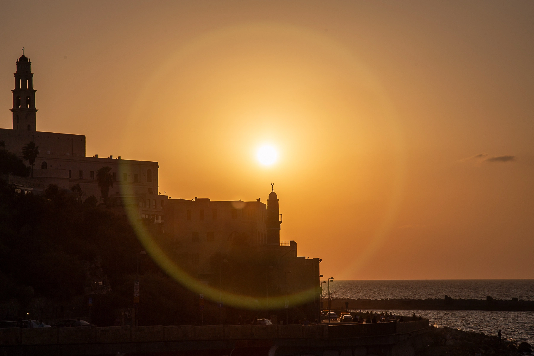 The sun sets over Jaffa Beach, casting a warm golden light on the old city and creating a dramatic lens flare in the foreground. Tel Aviv, Israel