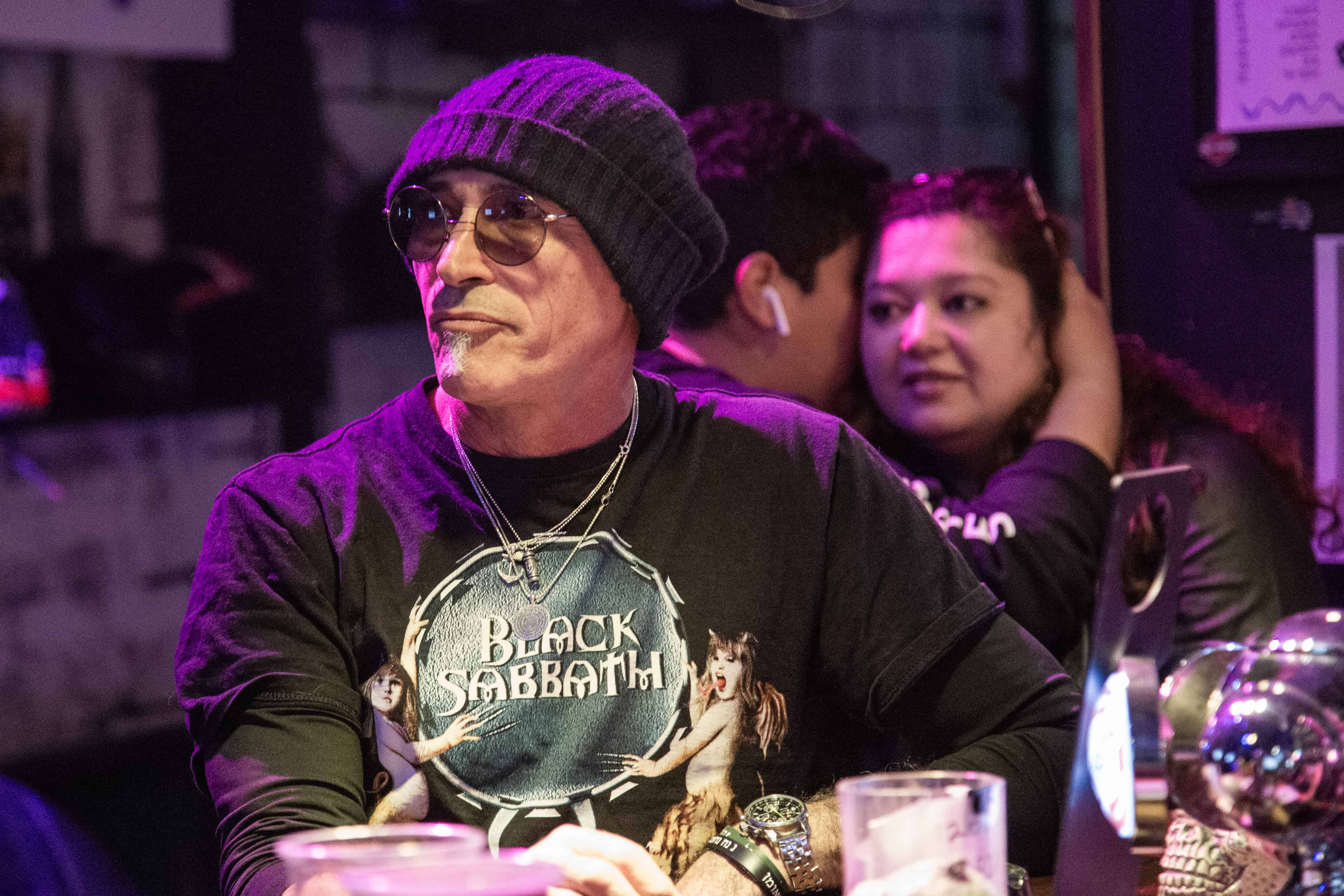 A man in a Black Sabbath shirt, sunglasses, and beanie enjoying a rock jam session in a bar. Tel Aviv, Israel