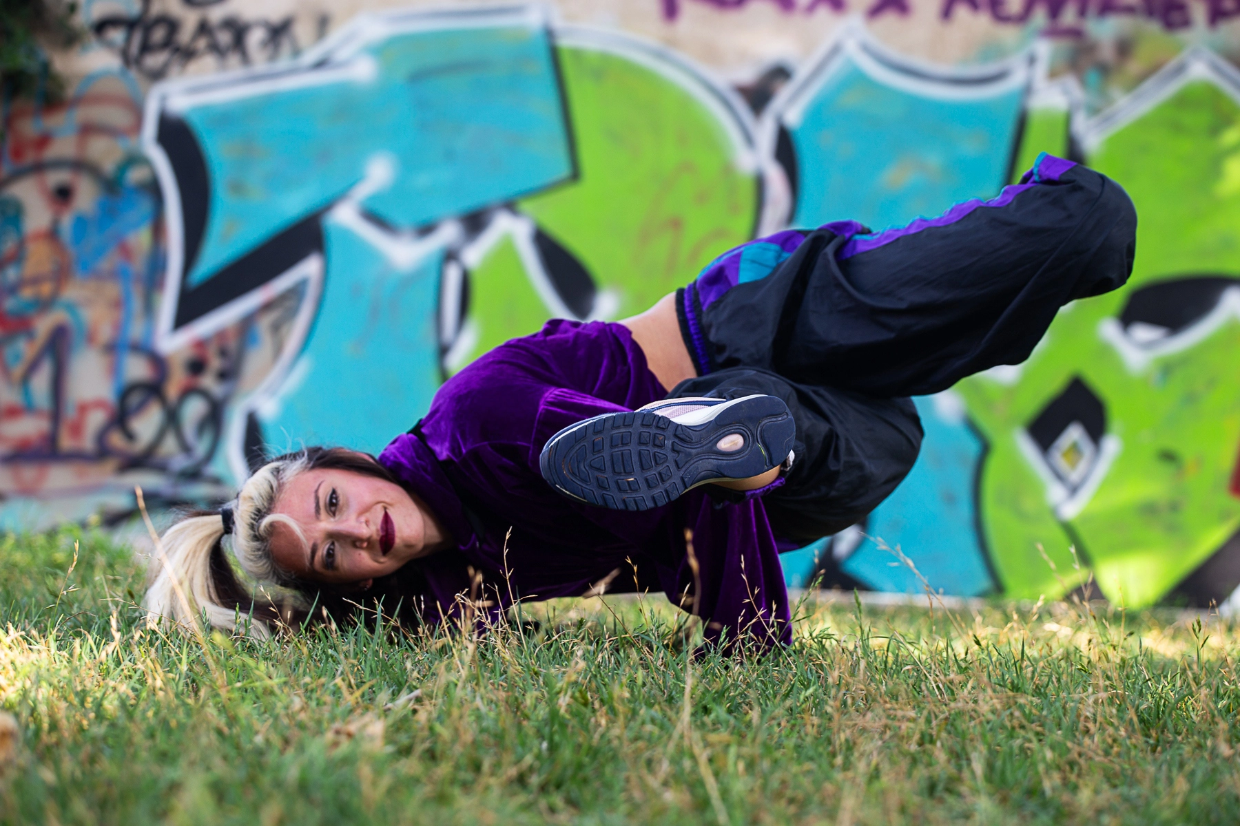 A woman performing a breakdance move on the grass, dressed in a purple outfit, with graffiti art in the background. Tel Aviv, Israel