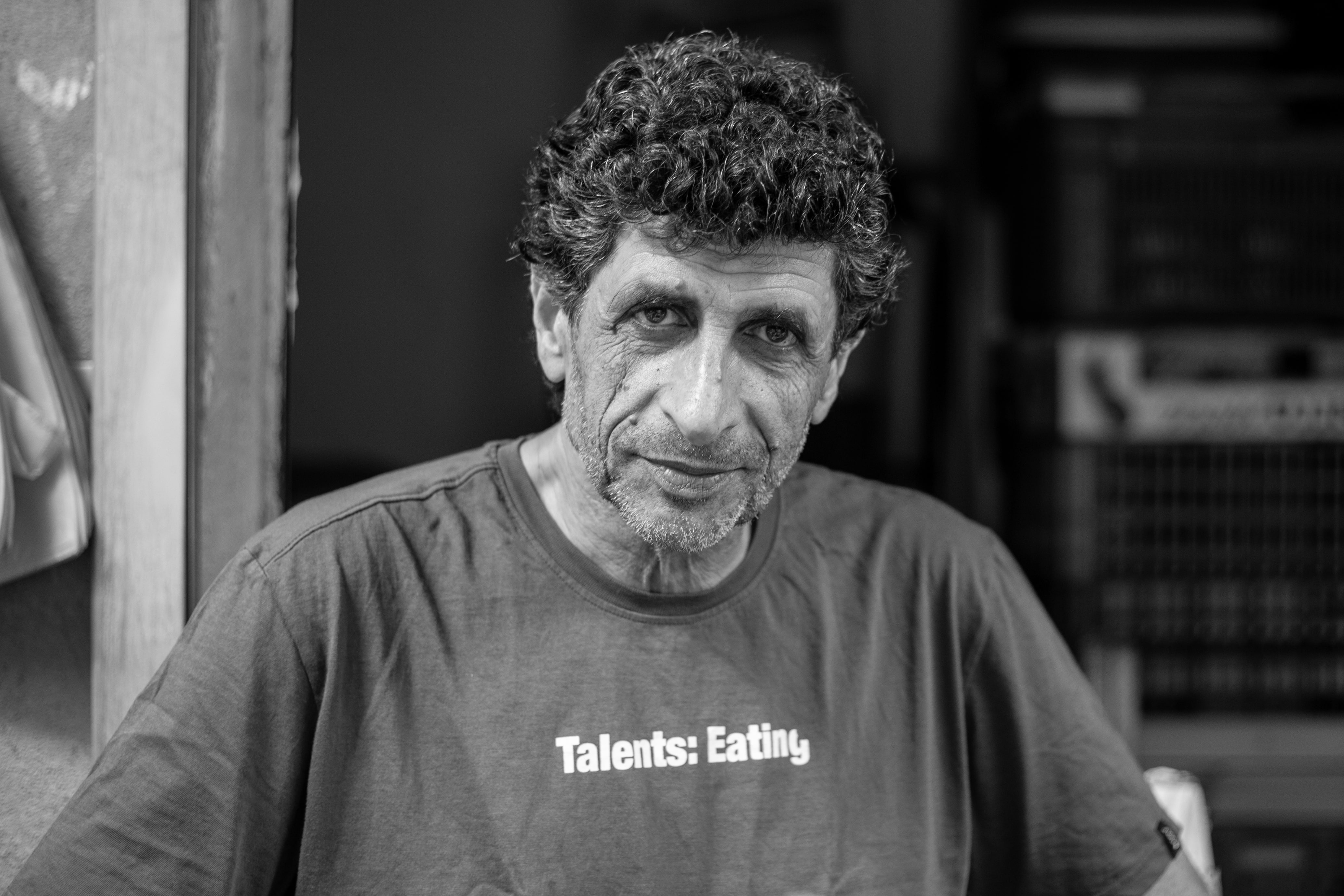 A black-and-white portrait of a man with curly hair, wearing a "Talents: Eating" shirt. Tel Aviv, Israel