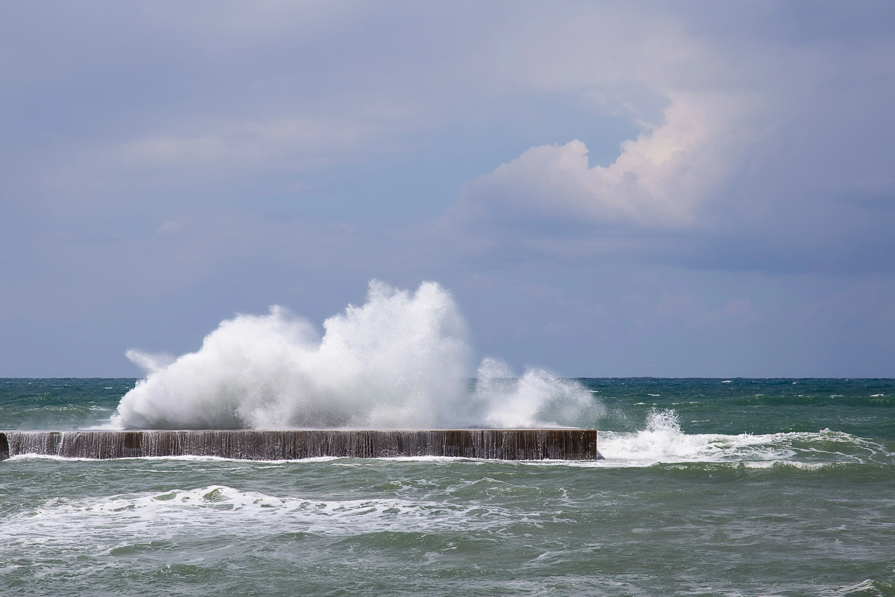 A massive wave crashes against a concrete structure in the ocean, with a cloudy sky above and turbulent waters below. Caesarea ,Israel