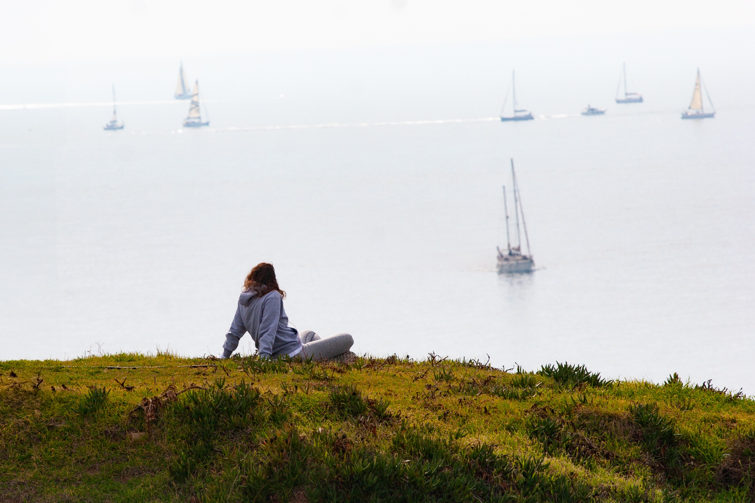 A person sits on a grassy hill overlooking the sea with several sailboats in the distance. Hertzliya, Israel