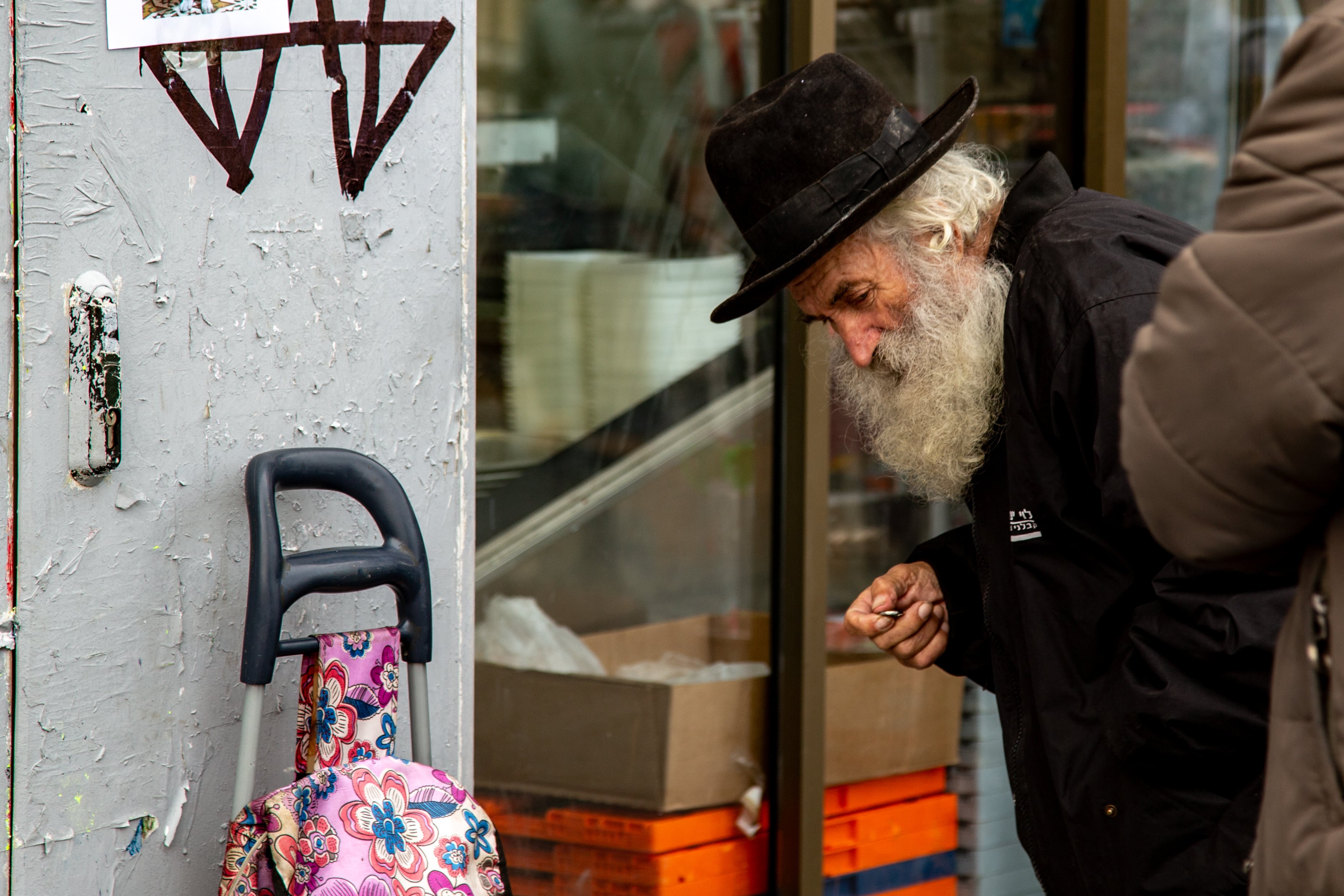 An elderly man holds a coin in his hand next to a bright floral cart in a muted urban setting in Jerusalem. Jerusalem, Israel