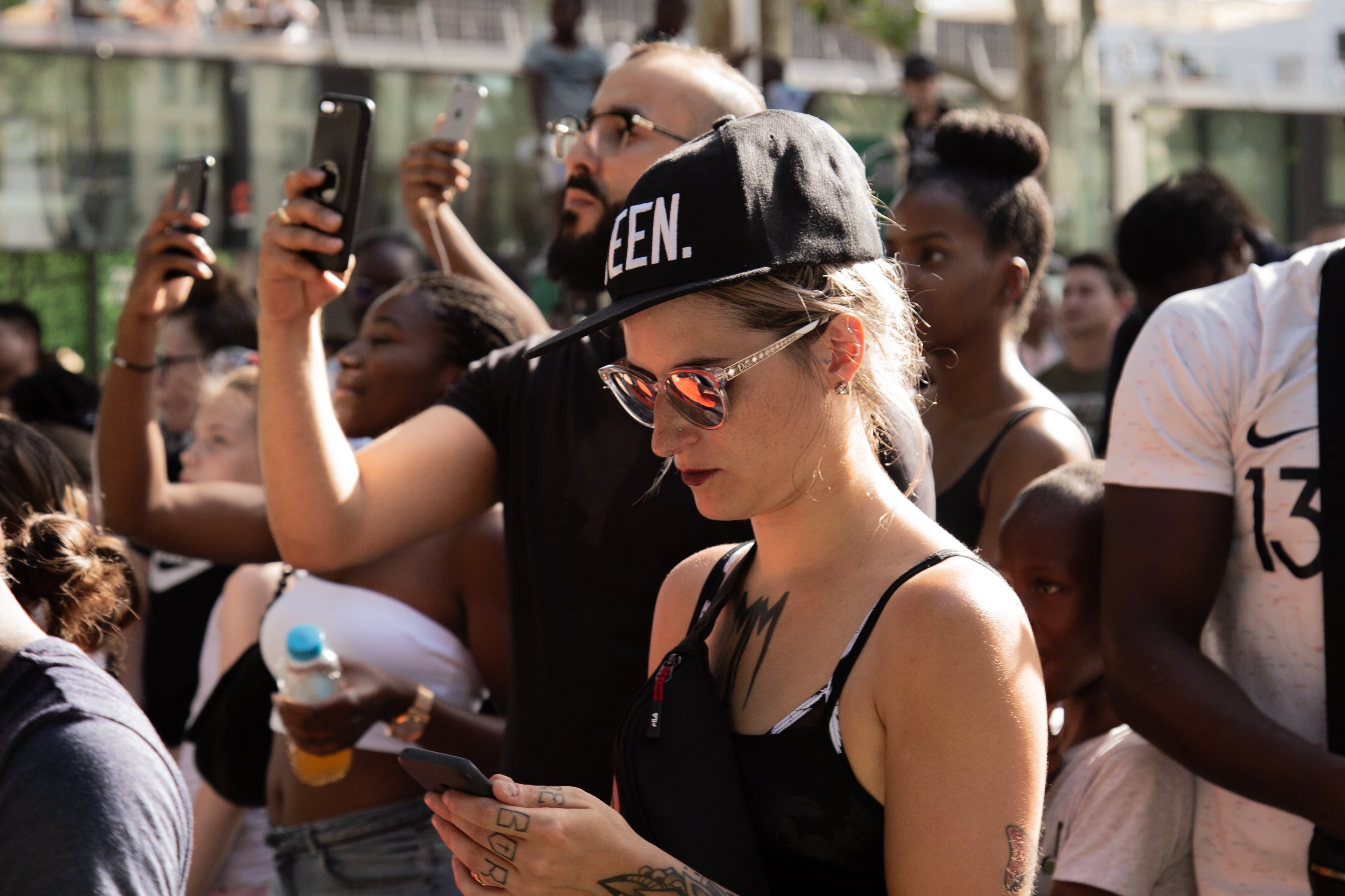 A woman in a “QUEEN” cap checks her phone at a concert, ignoring the crowd holding up phones around her. Paris, France