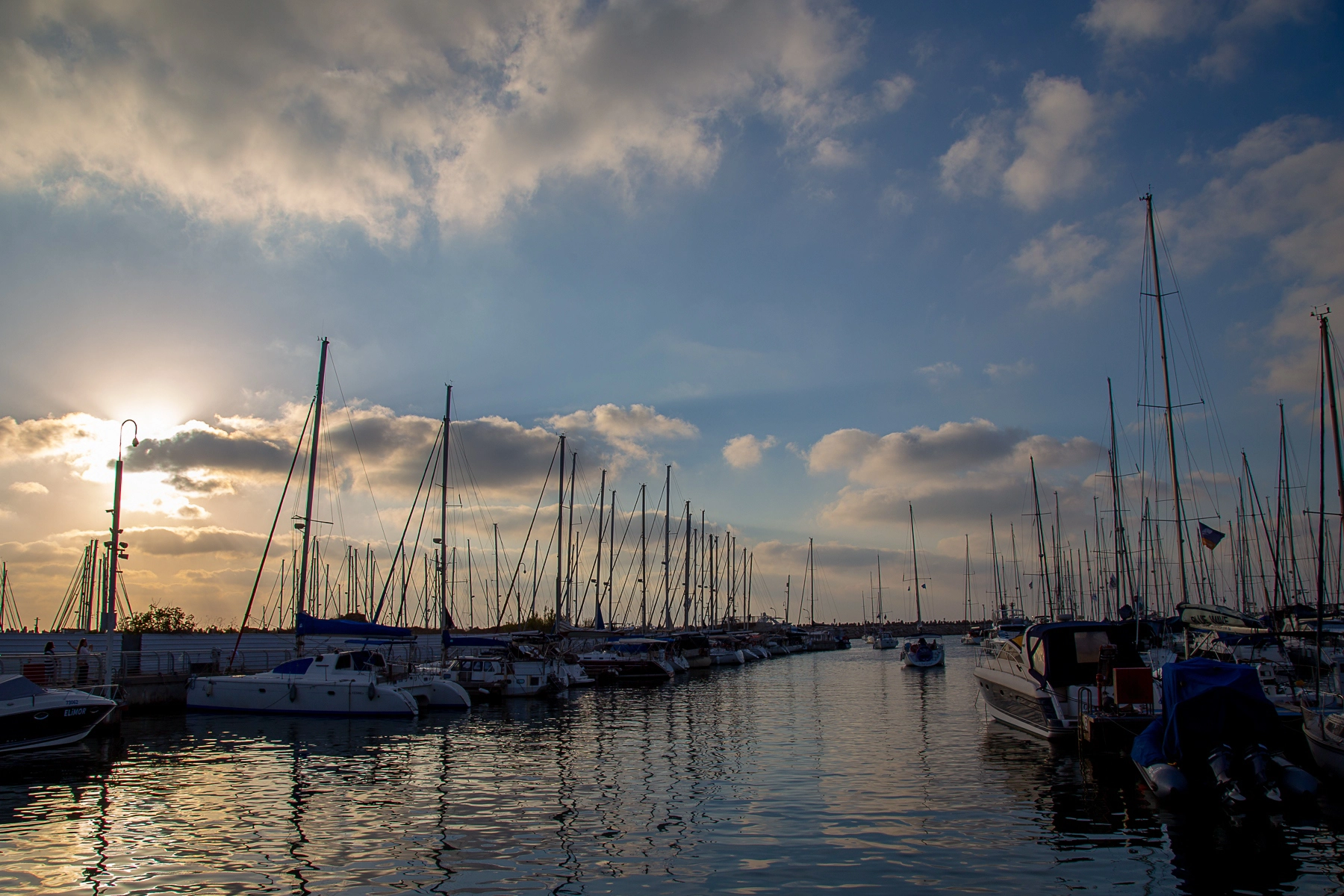 A tranquil view of boats docked at a marina, with the sun setting behind clouds and reflecting on the water. Hertzliya, Israel