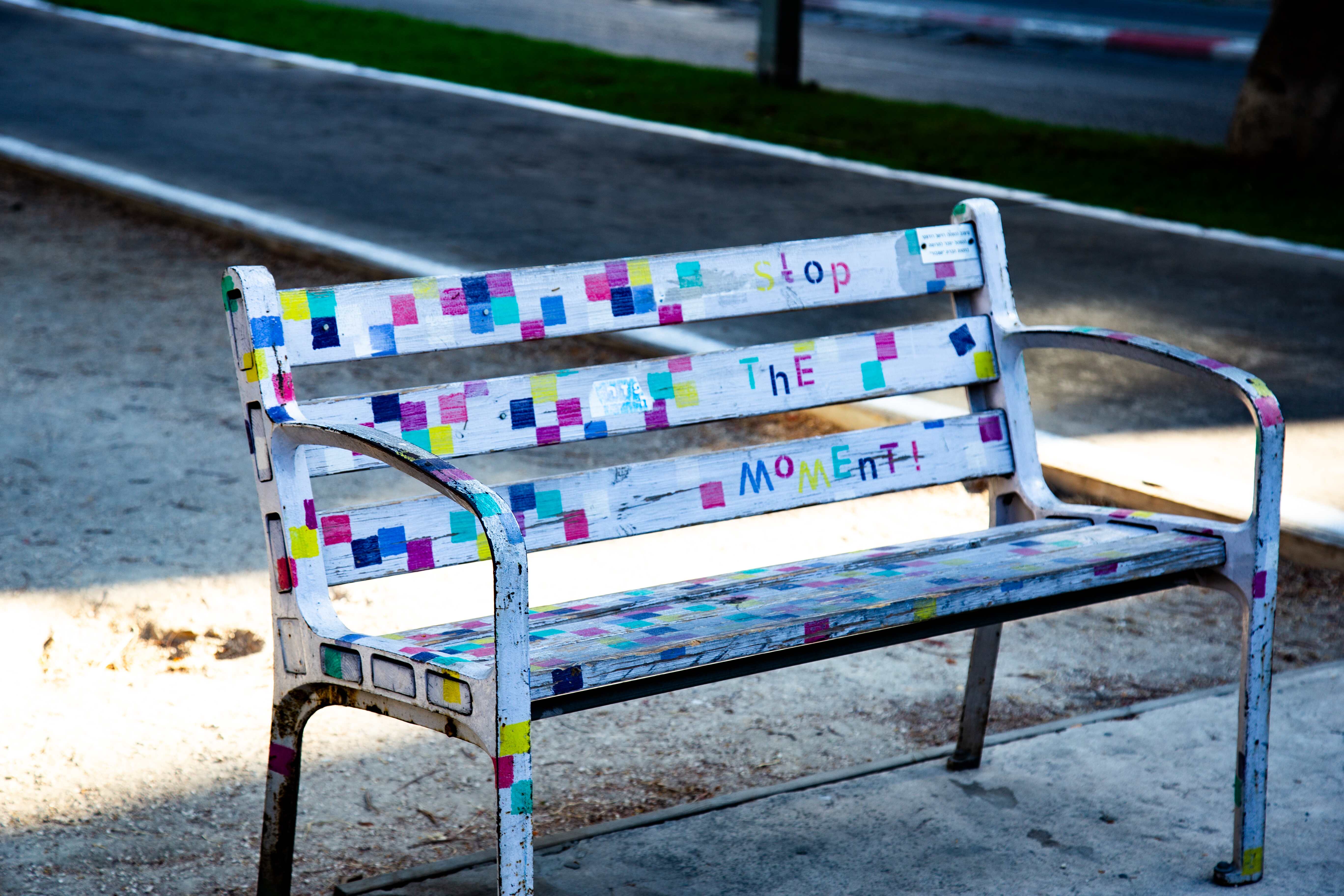 A white bench decorated with colorful squares and the words “Stop the Moment!” painted on its backrest. Tel Aviv, Israel