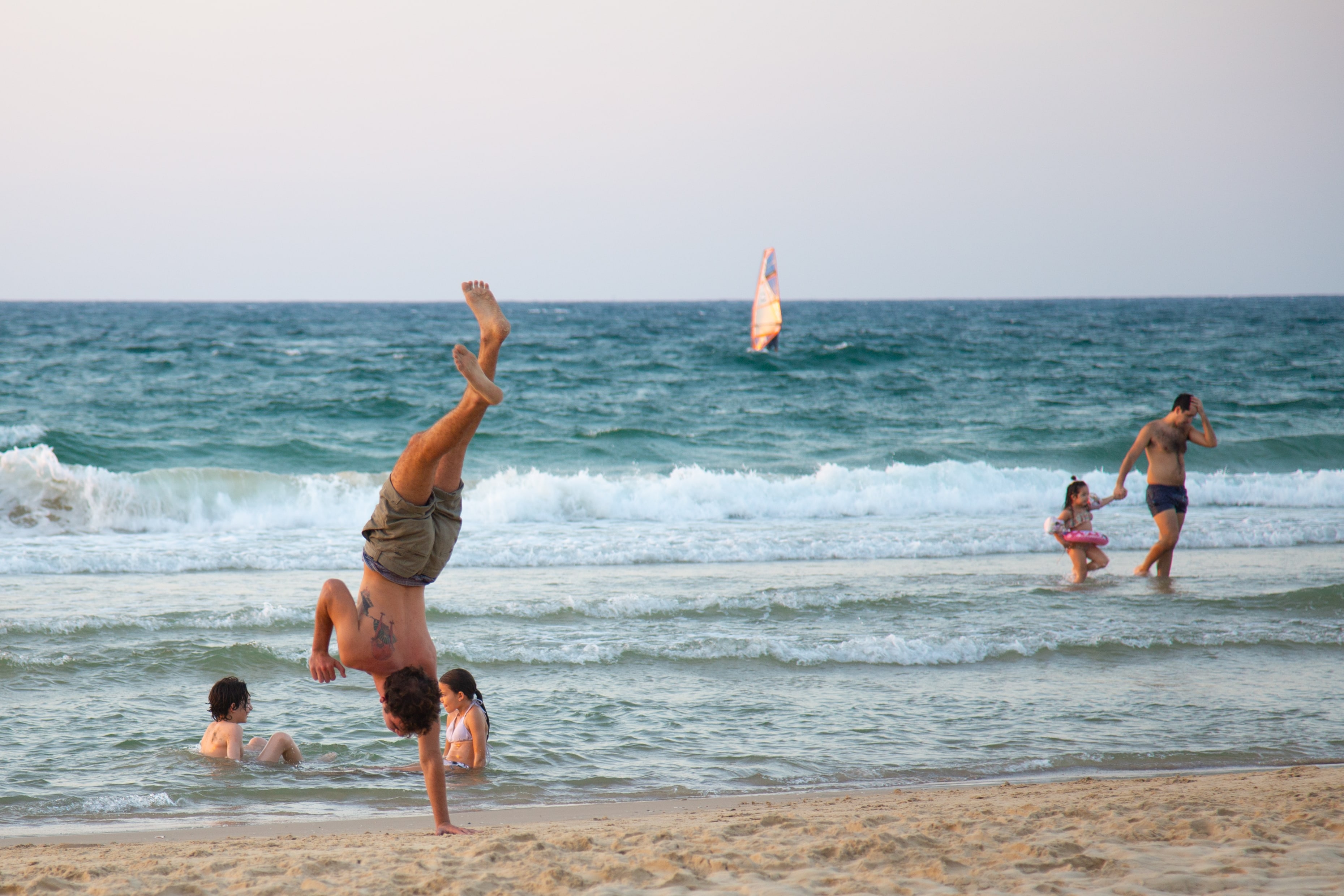 A man performs a handstand on Jaffa Beach as a windsurfer rides the waves in the background on a sunny day. Jaffa, Israel