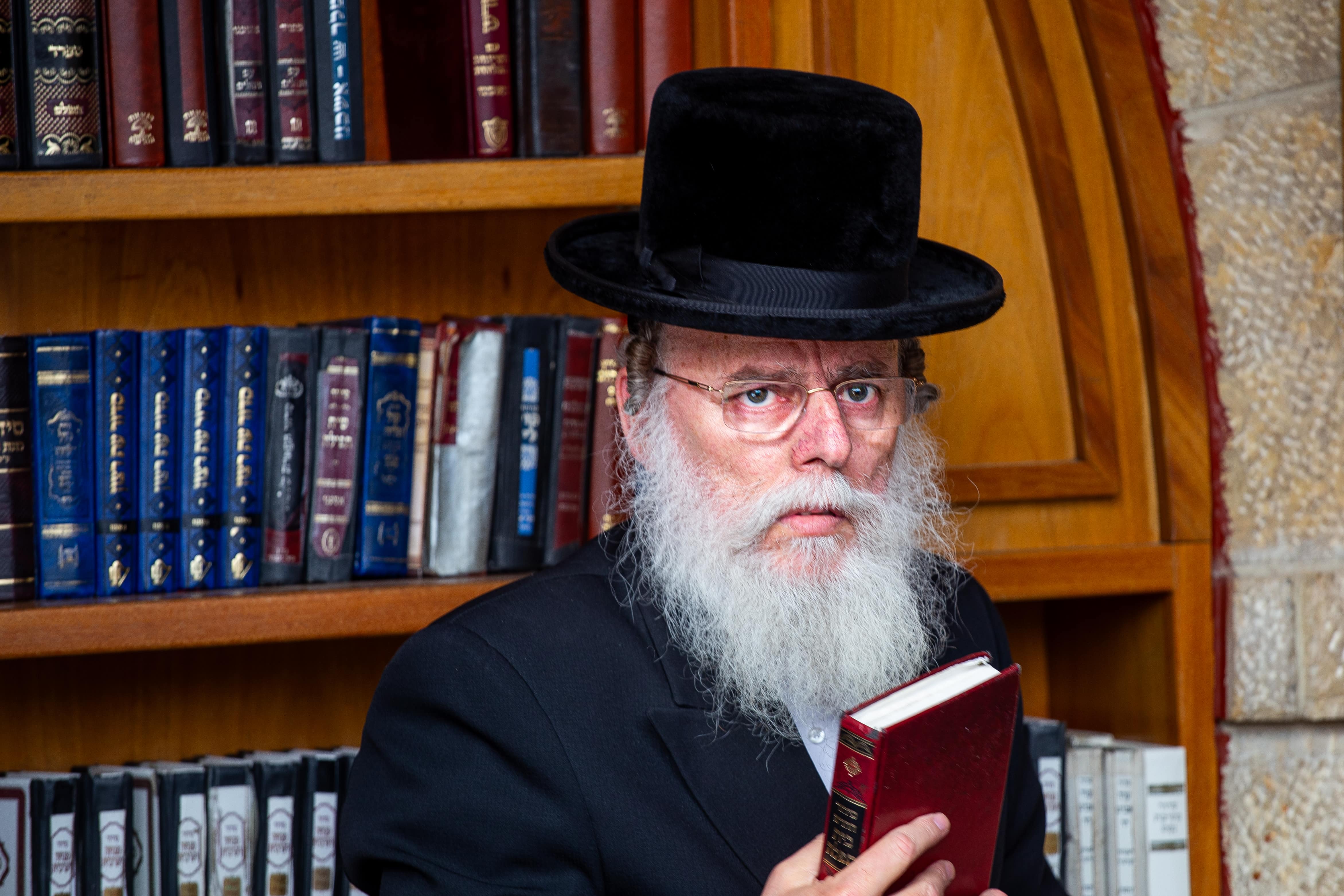 A bearded rabbi in traditional attire holding a book, standing in front of a bookshelf in Jerusalem's Old City.  Jerusalem, Israel