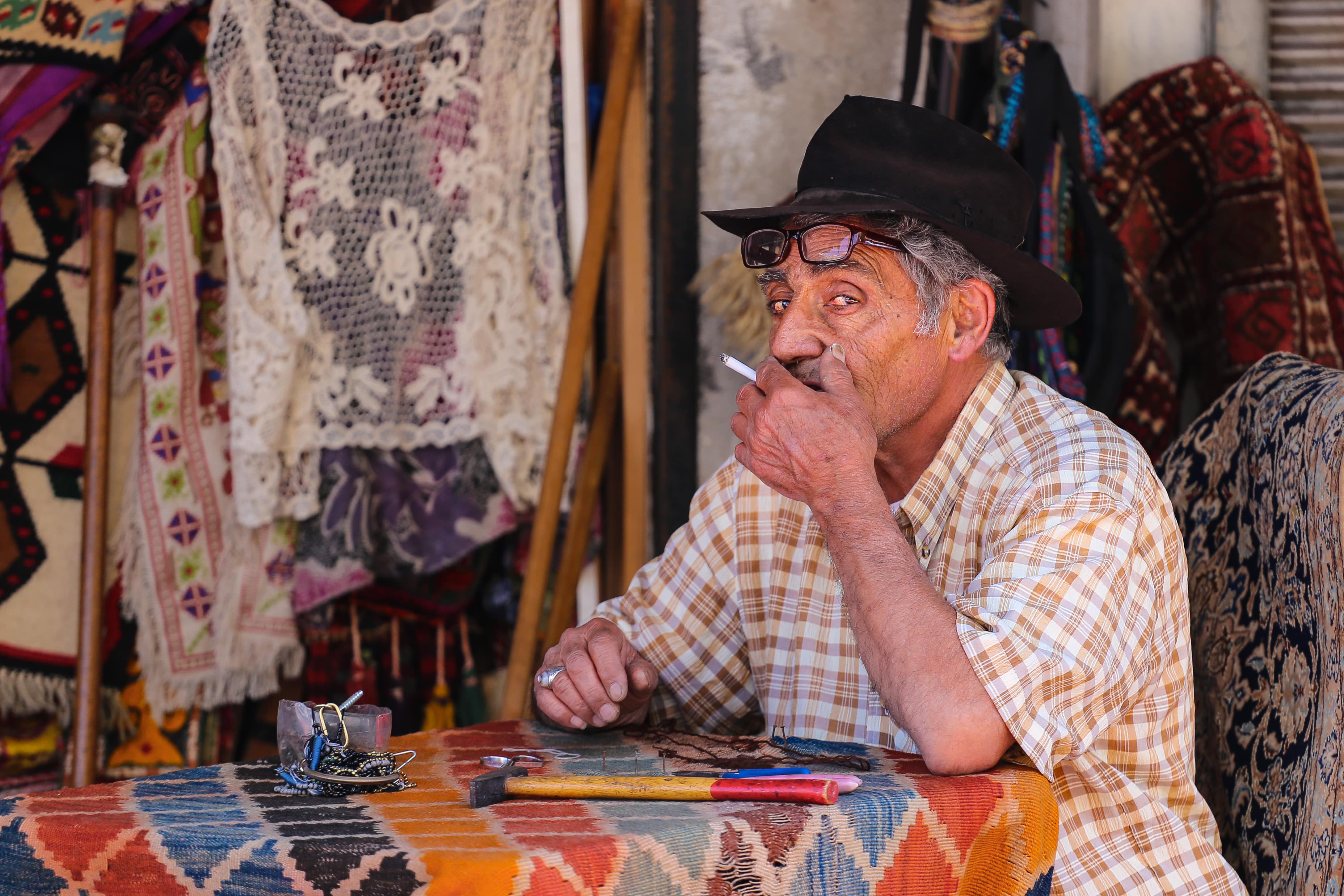 A man in a plaid shirt and hat, smoking a cigarette, sits at a table covered in fabrics, surrounded by handmade goods. Jaffa Flea Market,Israel