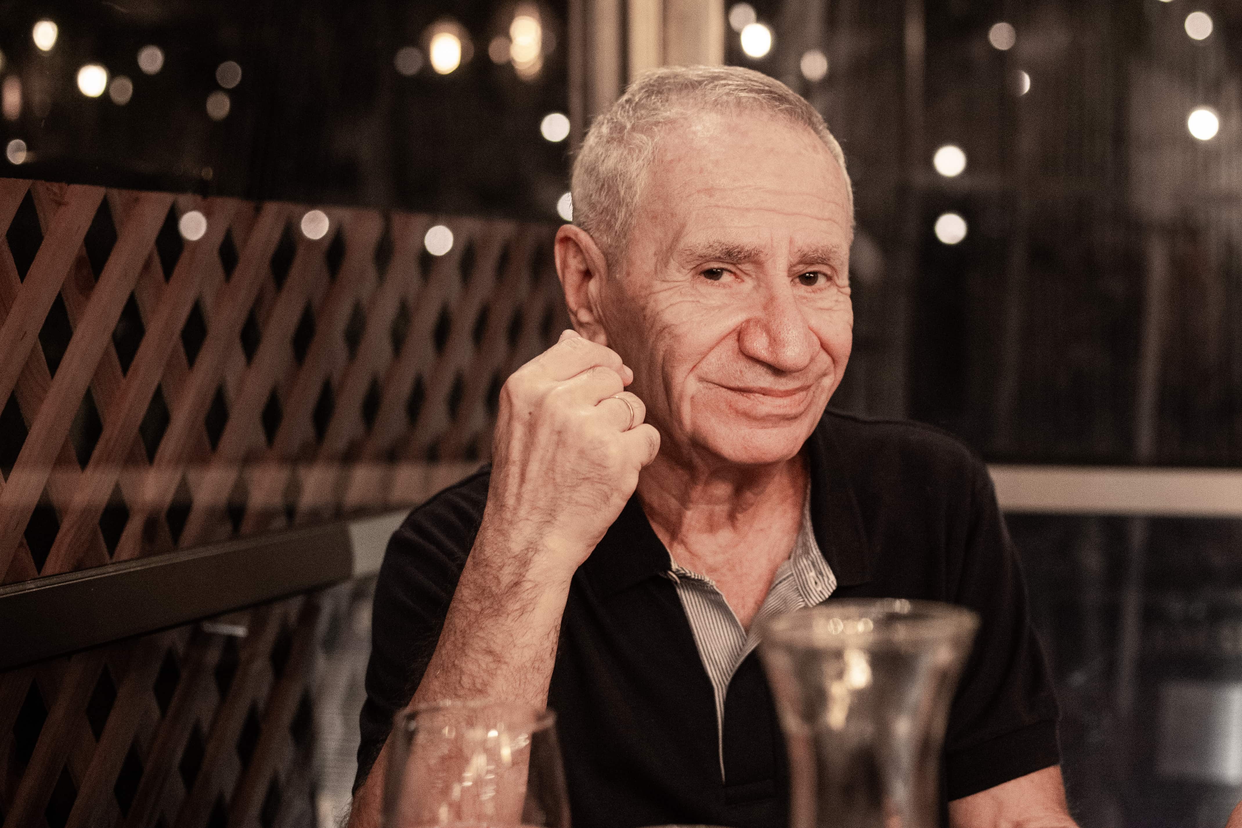 A man with a slight smile sits at a table in a warmly lit setting, his hand gently raised. Tel Aviv, Israel