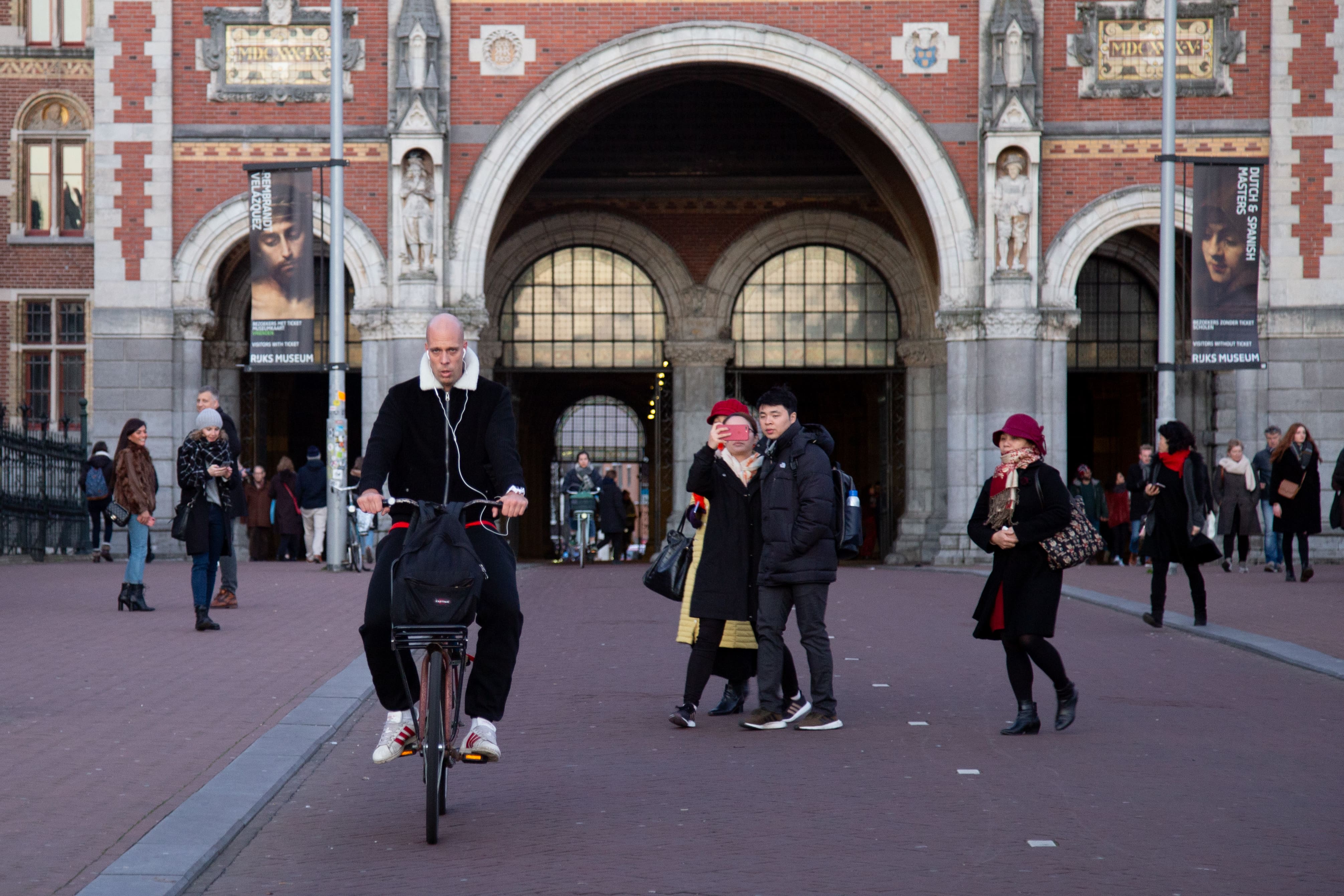 A cyclist rides past the Rijksmuseum courtyard, while tourists stroll and capture the lively scene. Rijksmuseum, Amsterdam, Netherlands