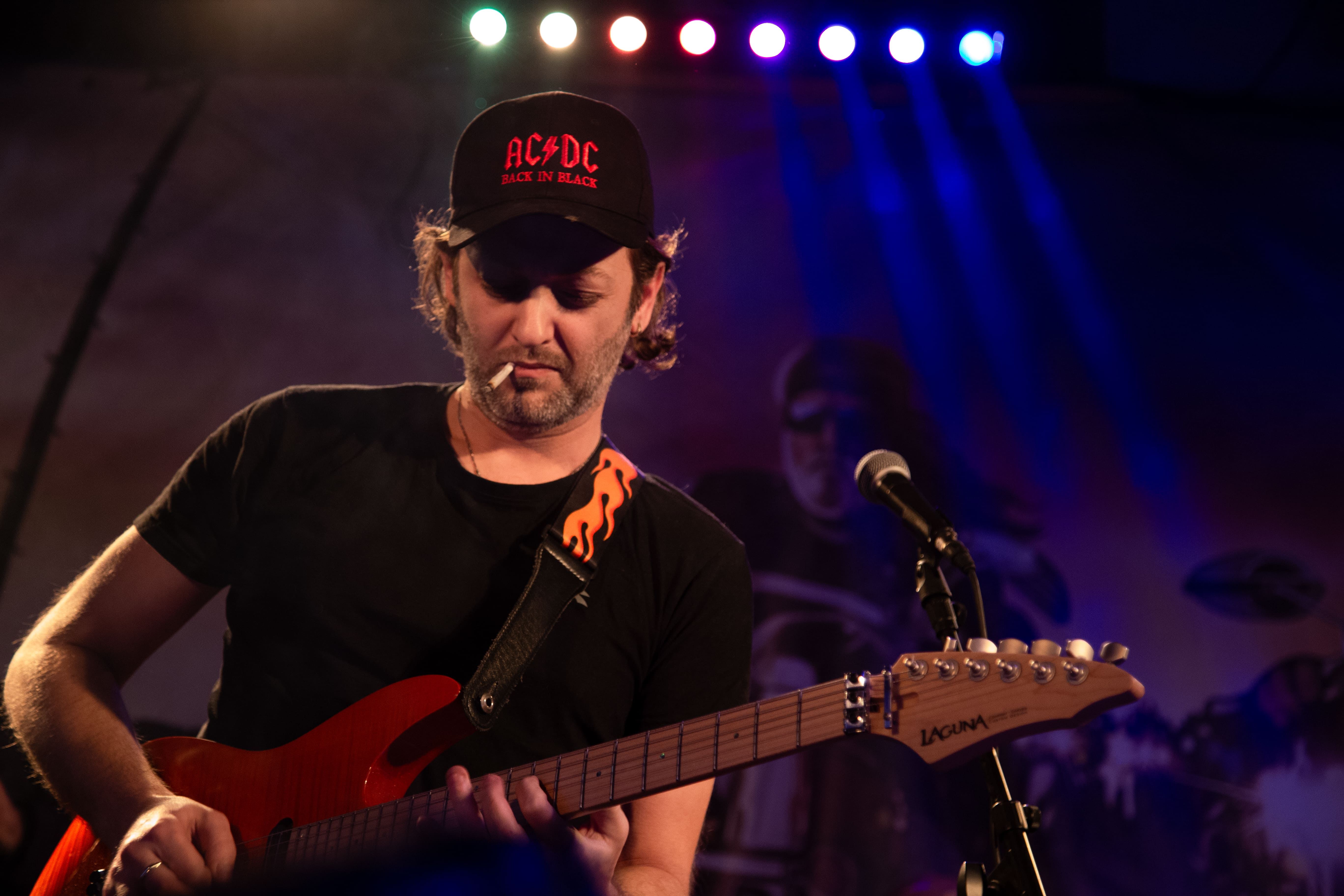 A guitarist in an AC/DC cap plays an electric guitar during a live performance, colorful stage lights in the background. Garage Pub, Israel 