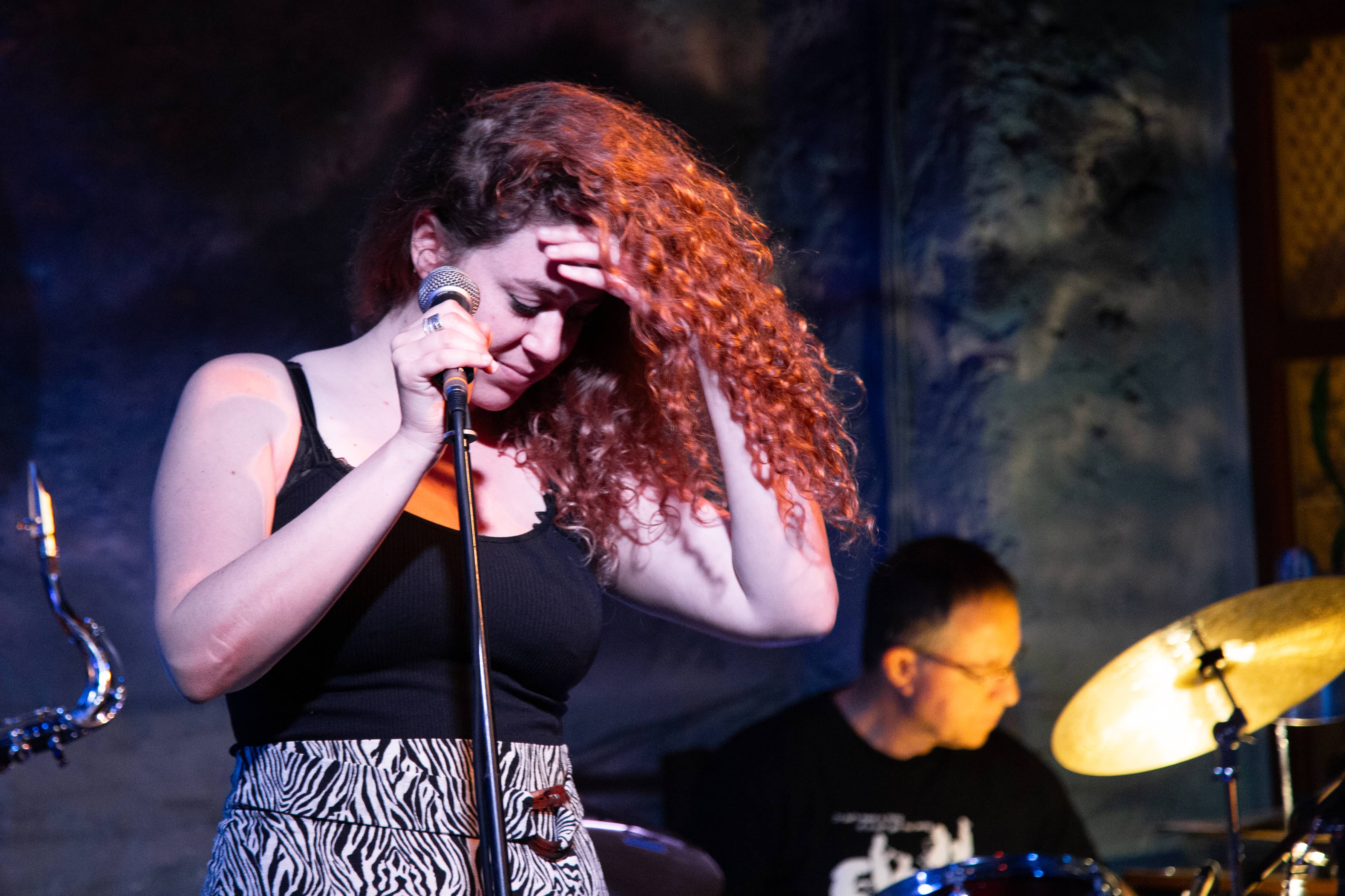 A woman with curly hair sings into a microphone, eyes closed, while a drummer plays behind her. Tel Aviv, Israel 