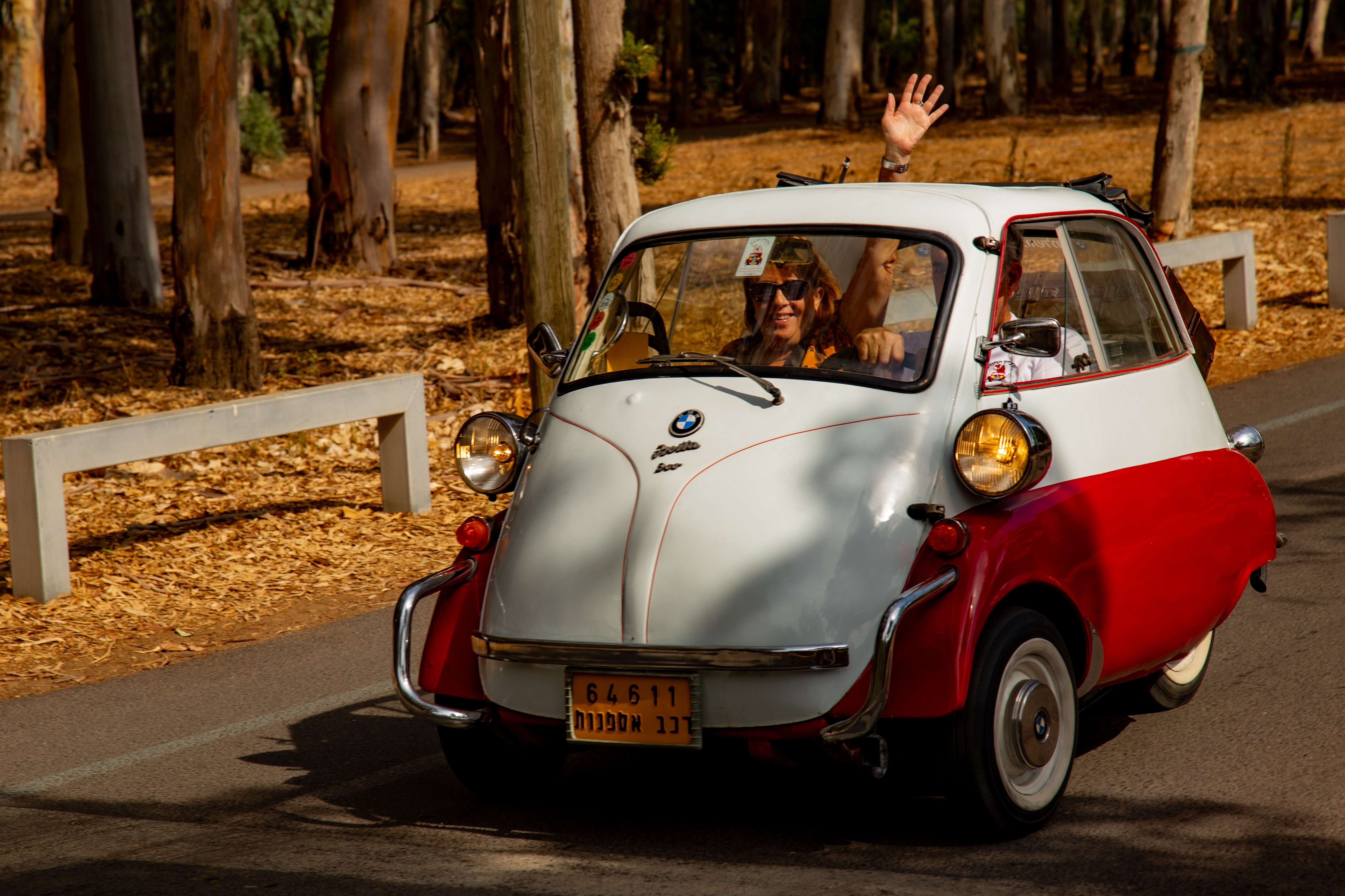 A cheerful driver in a vintage red and white BMW Isetta, waving while driving through a tree-lined park road. Retro Car Club Leumi, Tel Aviv, Israel