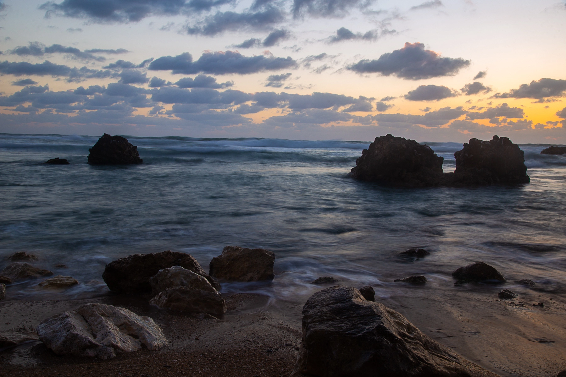 A peaceful beach scene at twilight with waves rolling in, rocks scattered on the shore, and soft light fading behind clouds. Sidney Ali Beach, Israel