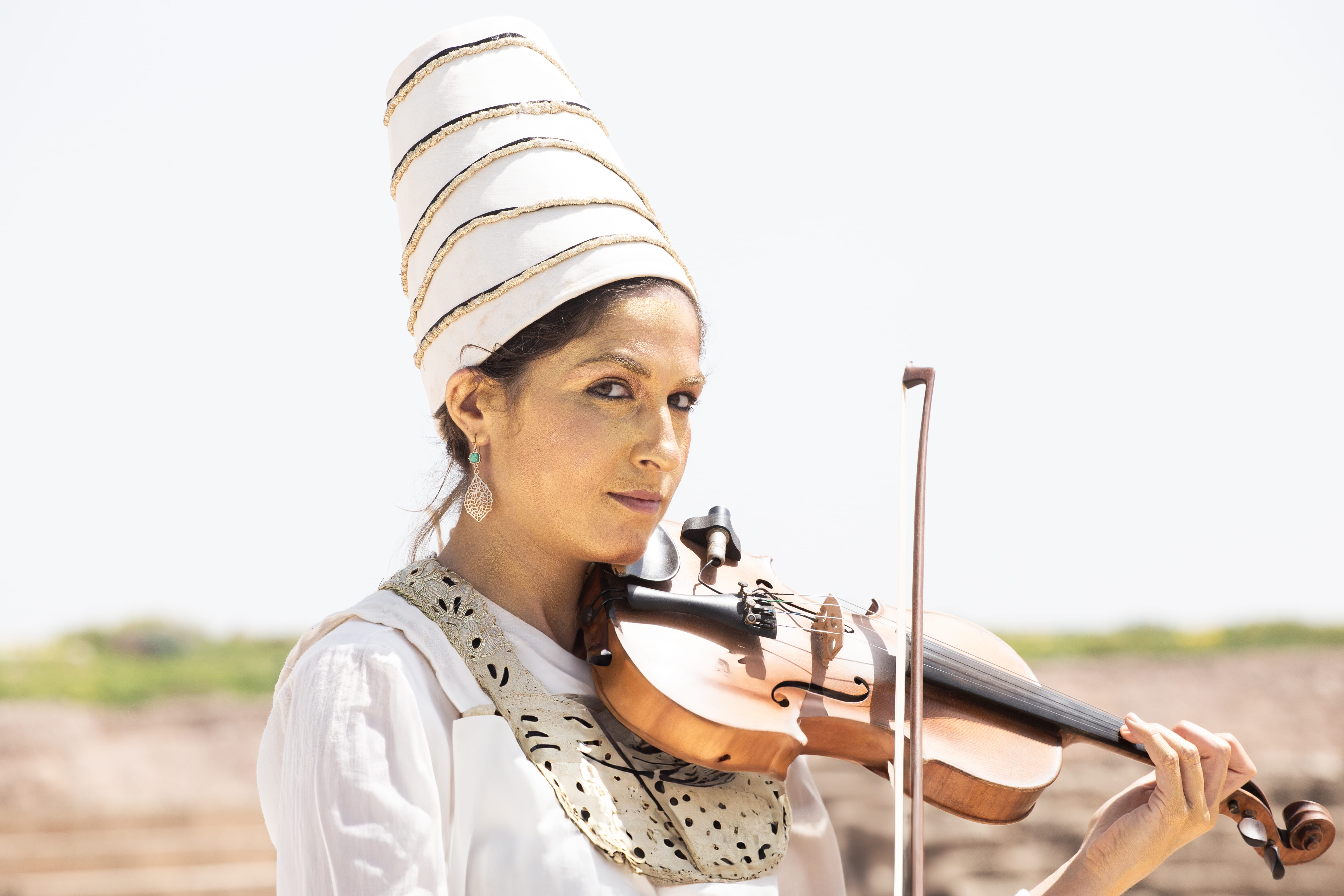 A woman wearing traditional white attire and a tall headdress plays the violin outdoors. Caesarea, Israel