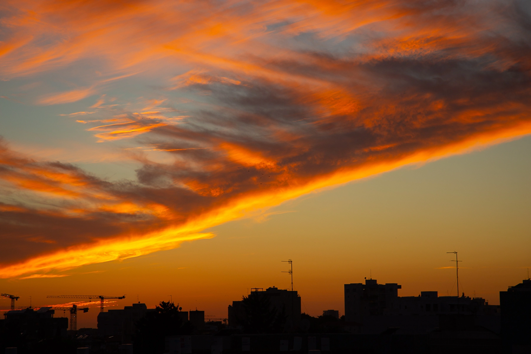 A vibrant sunset with streaks of orange and red clouds cutting across the sky, with the silhouettes of buildings and cranes below. Hertzliya, Israel