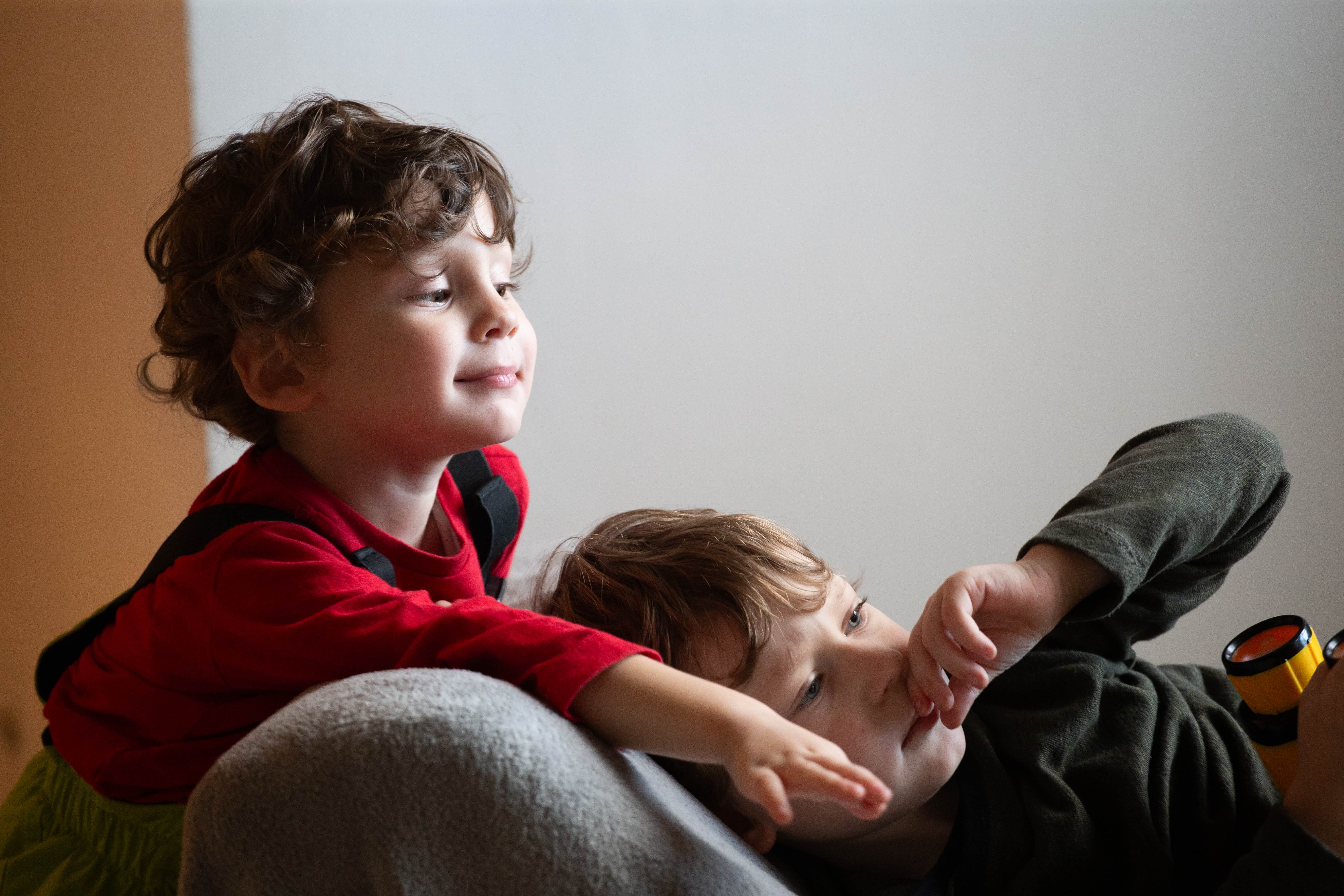 Two young boys, one smiling and reaching out, the other relaxed and thoughtful, lying on a couch. Tel Aviv, Israel 