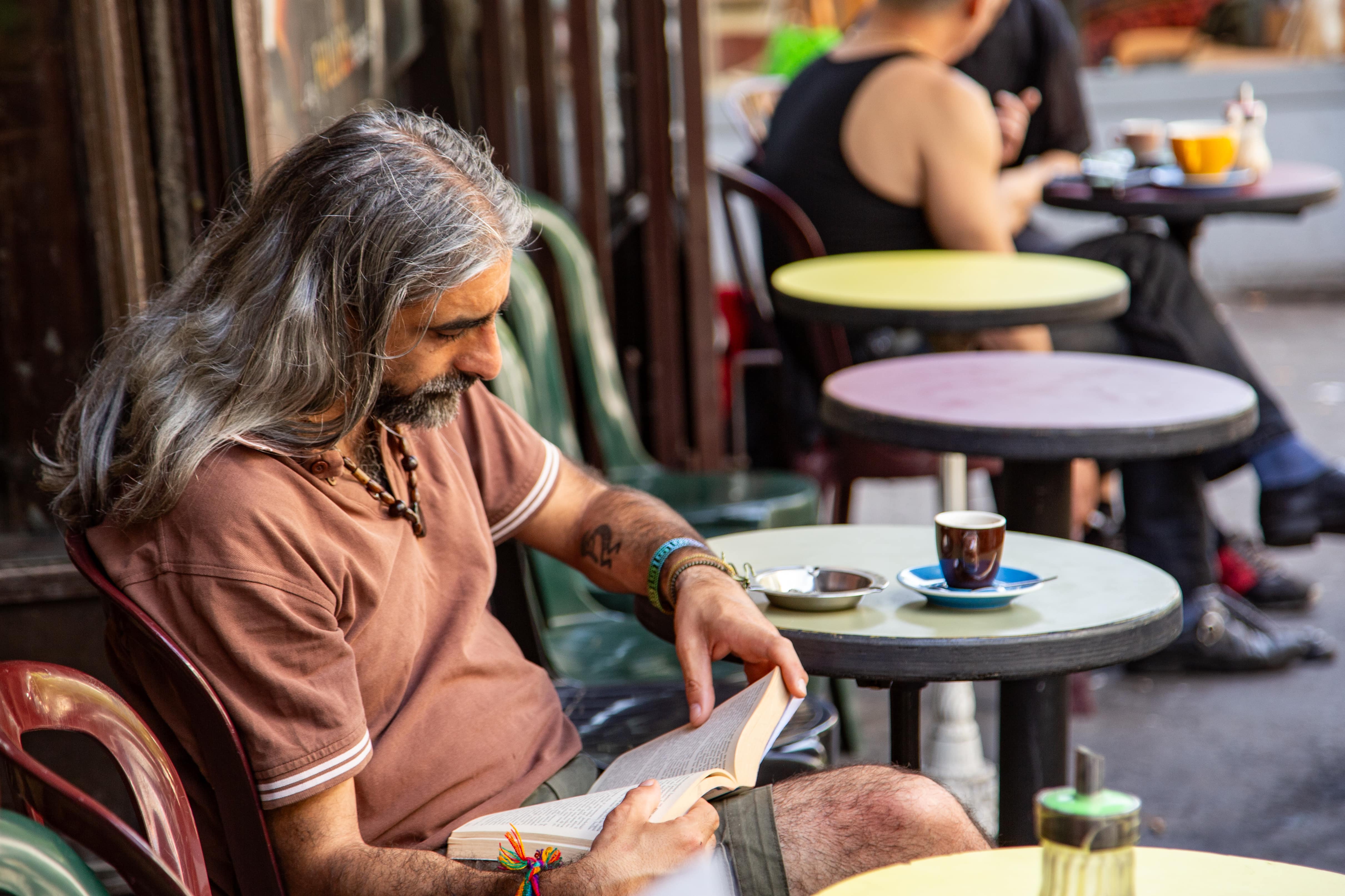 A man with long hair reads a book at an outdoor café table, enjoying a quiet moment with coffee. Paris, France