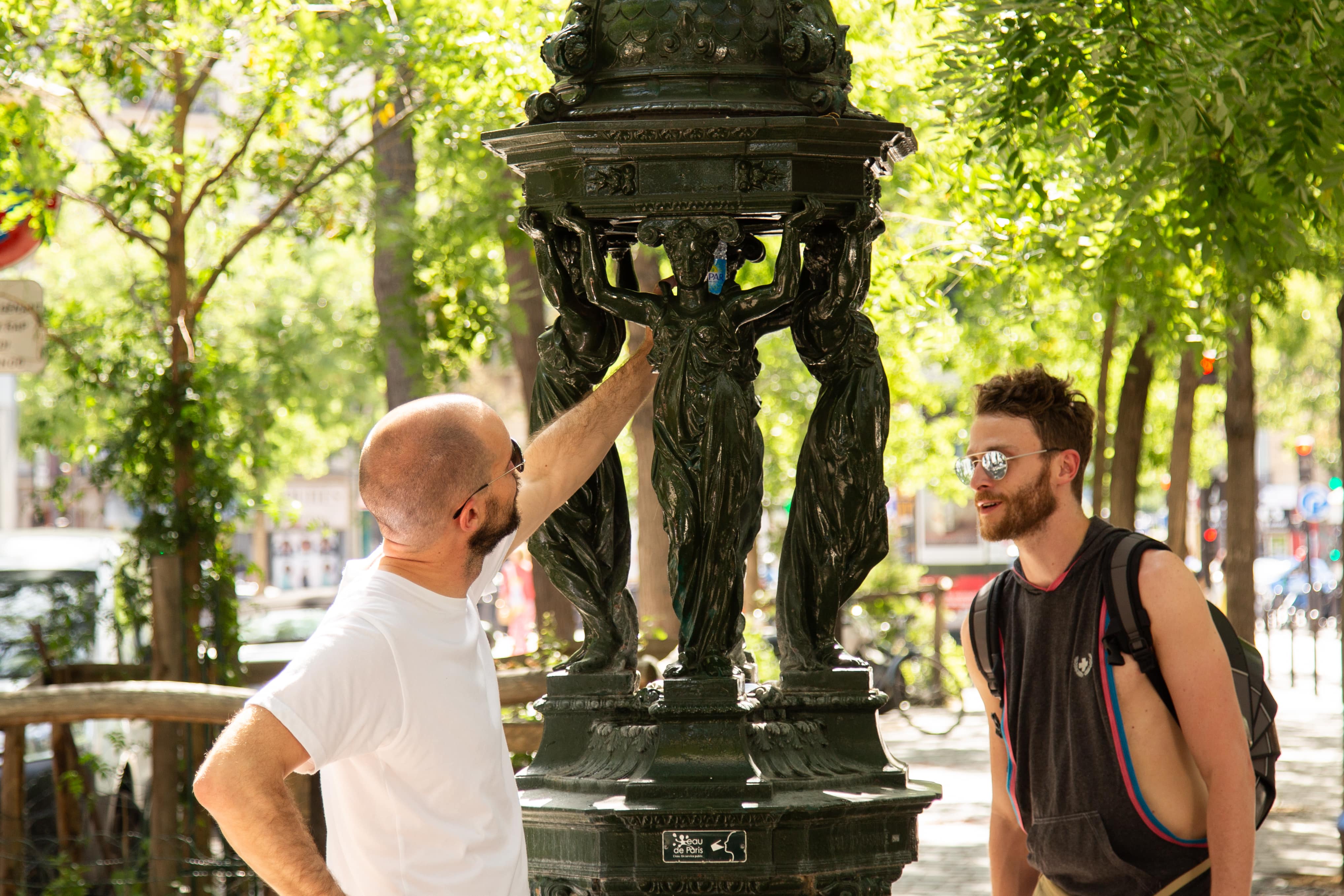 Two men stop to admire a street sculpture on a leafy Paris avenue, surrounded by greenery and outdoor art. Paris, France