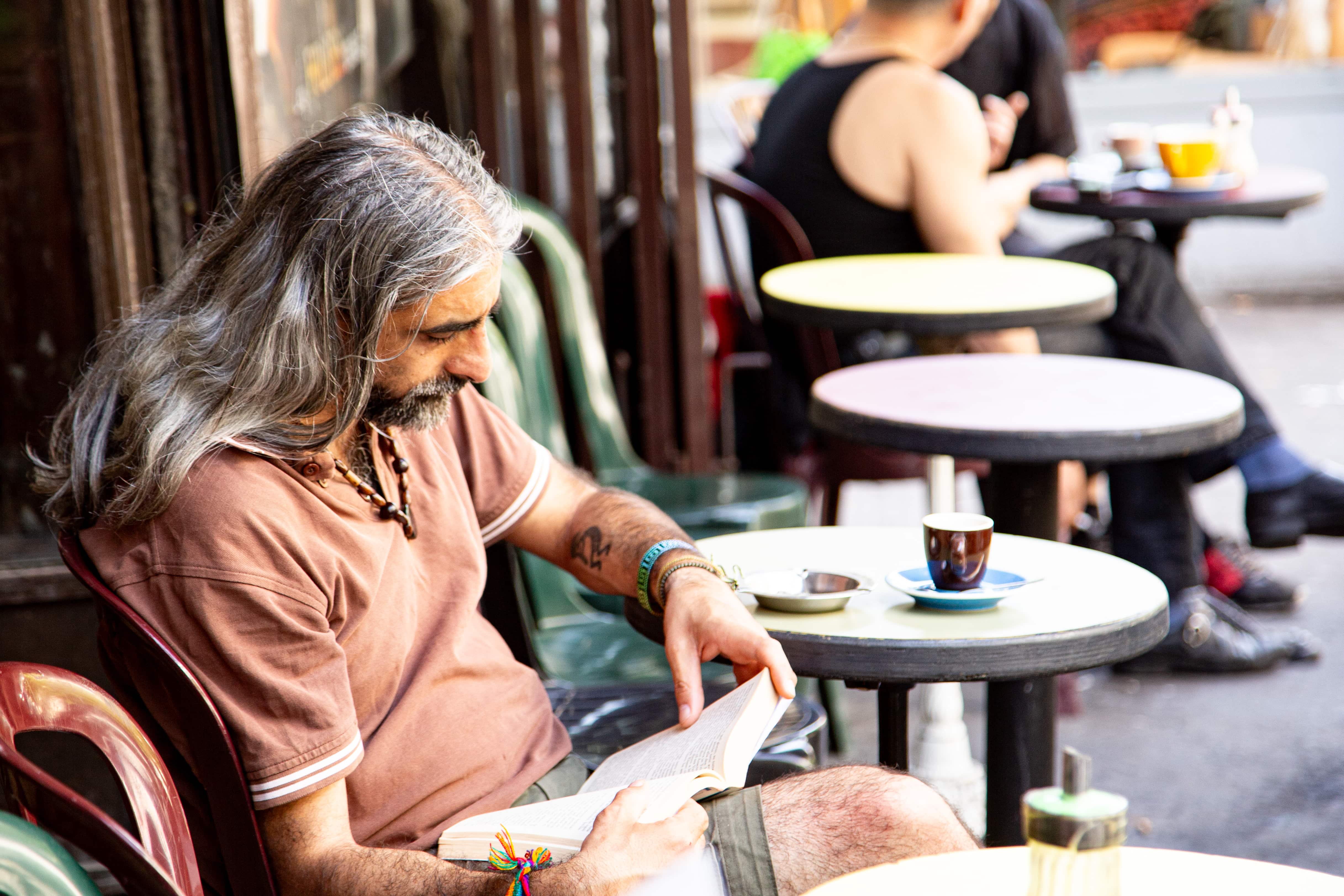 A man sits outside a Paris café, absorbed in his book, with a cup of coffee and vibrant street life around. Paris, France