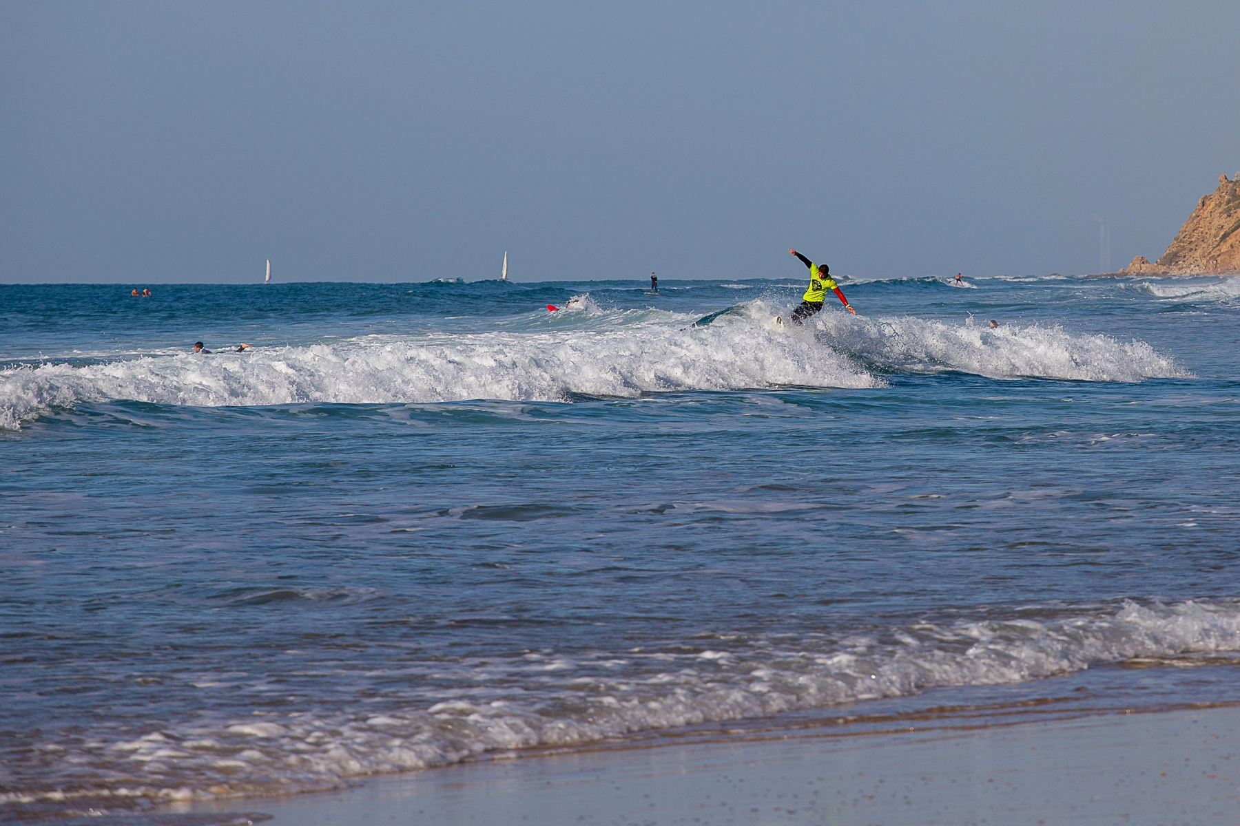 A surfer wearing a bright shirt catching a small wave with sailboats visible in the background. Herzliya, Israel