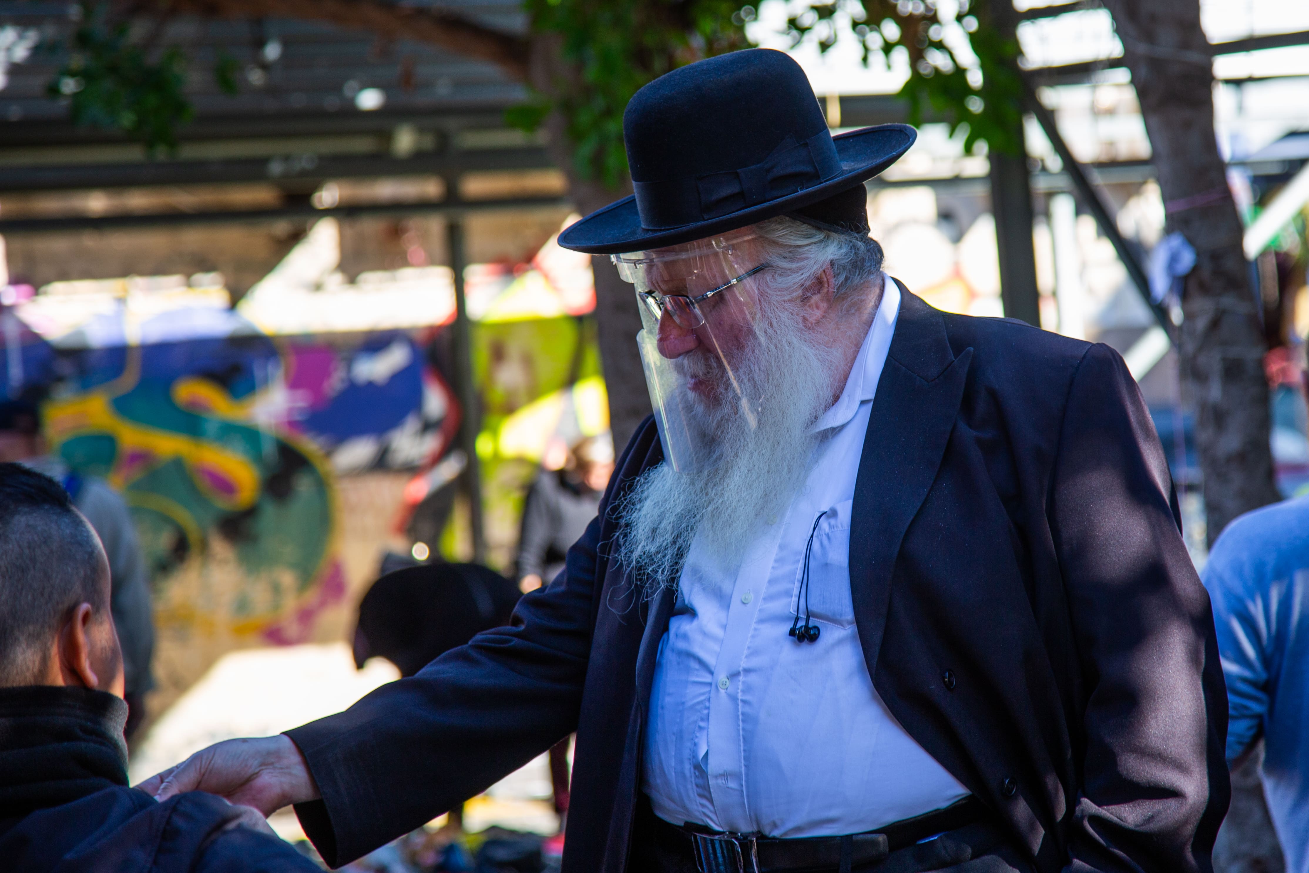 An Orthodox Jew with a face shield converses at Tel Aviv Flea Market, blending tradition with urban graffiti. Tel Aviv Flea Market, Israel