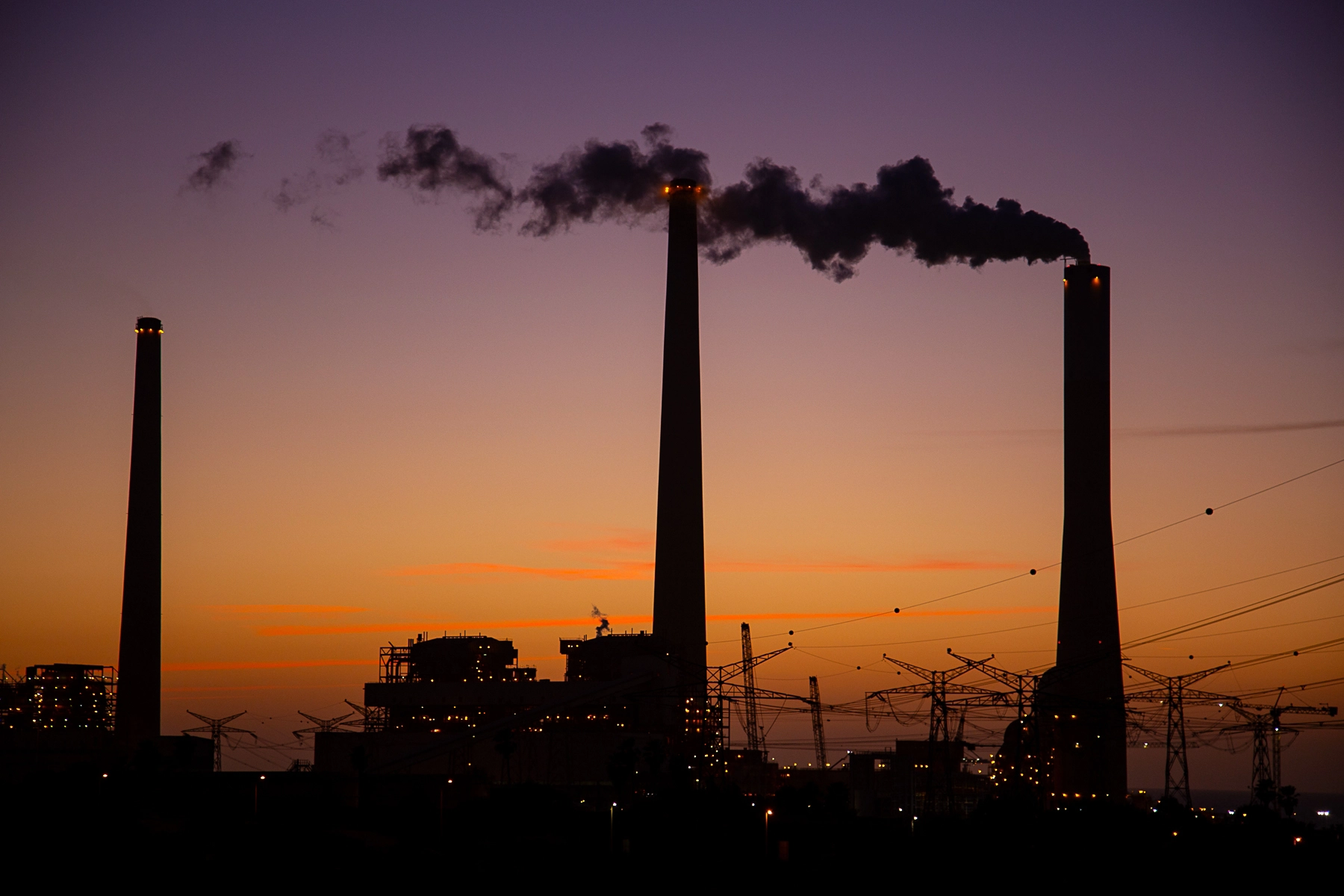 Tall smokestacks silhouetted against a purple and orange sunset, with dark smoke plumes rising into the sky. Hadera, Israel