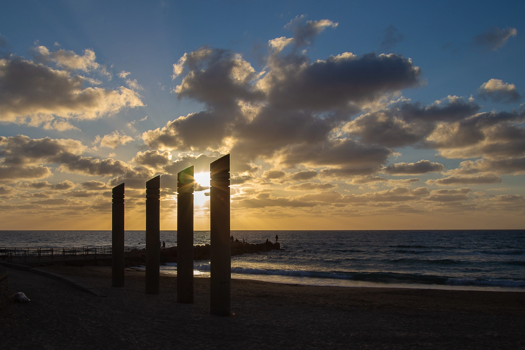 A peaceful beach scene with golden light shining through four standing pillars, casting long shadows and illuminating scattered clouds. Haifa, Israel