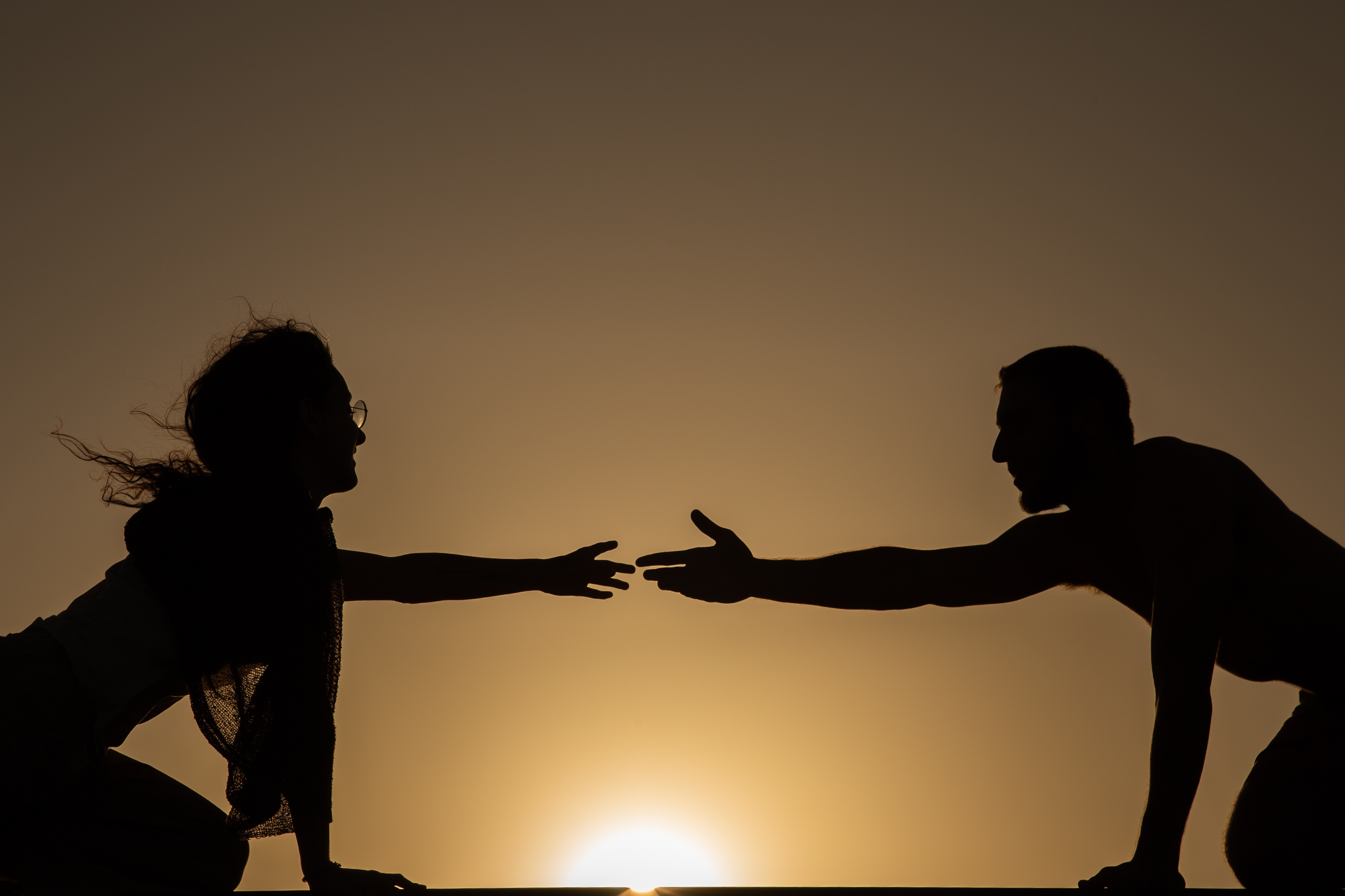 A dramatic silhouette of two people reaching out towards each other against a sunset backdrop. Mitzpe Ramon, Israel