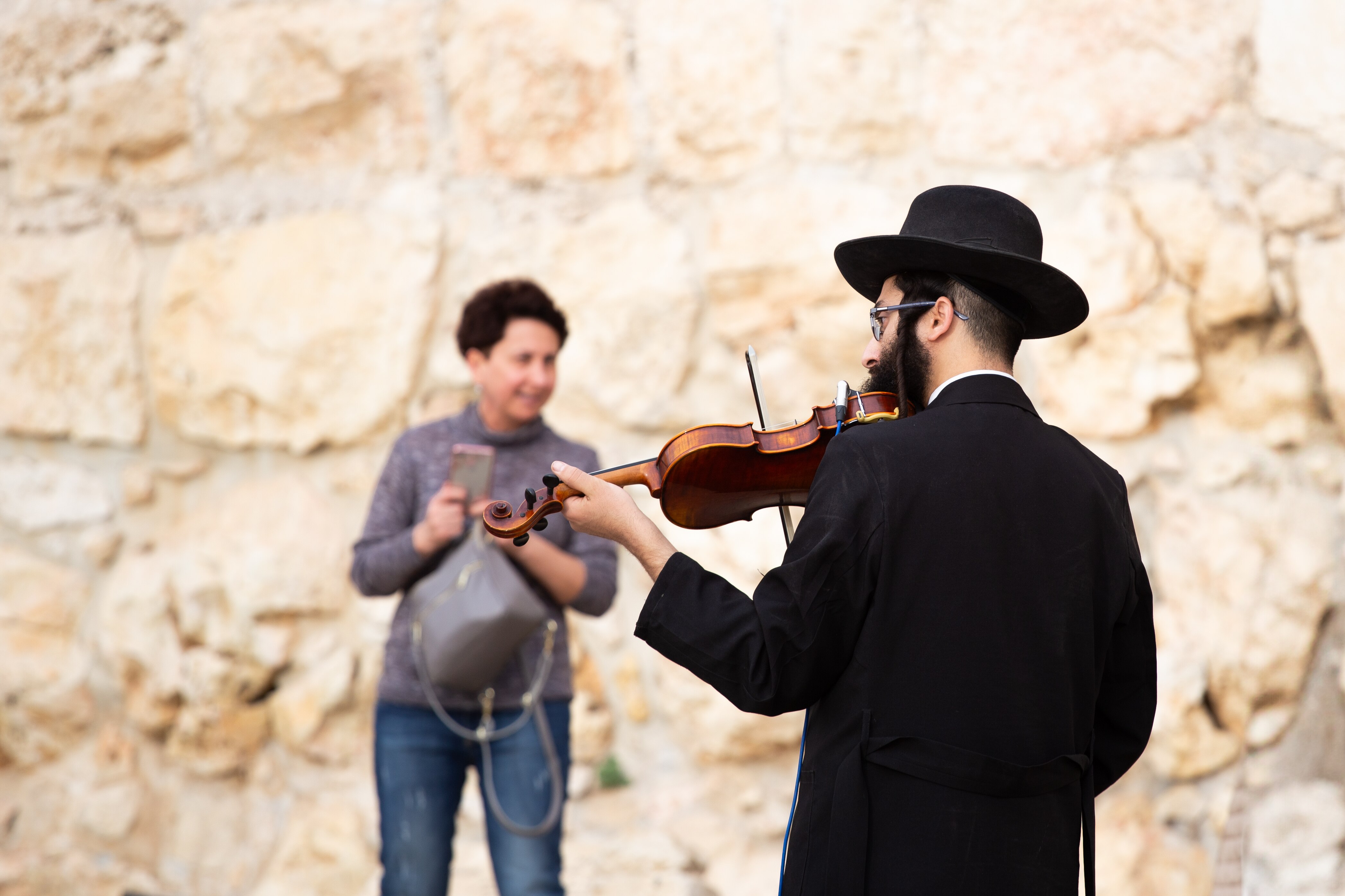 A violinist in traditional dress plays in front of a stone wall, captivating a nearby woman. Jerusalem Old City, Israel