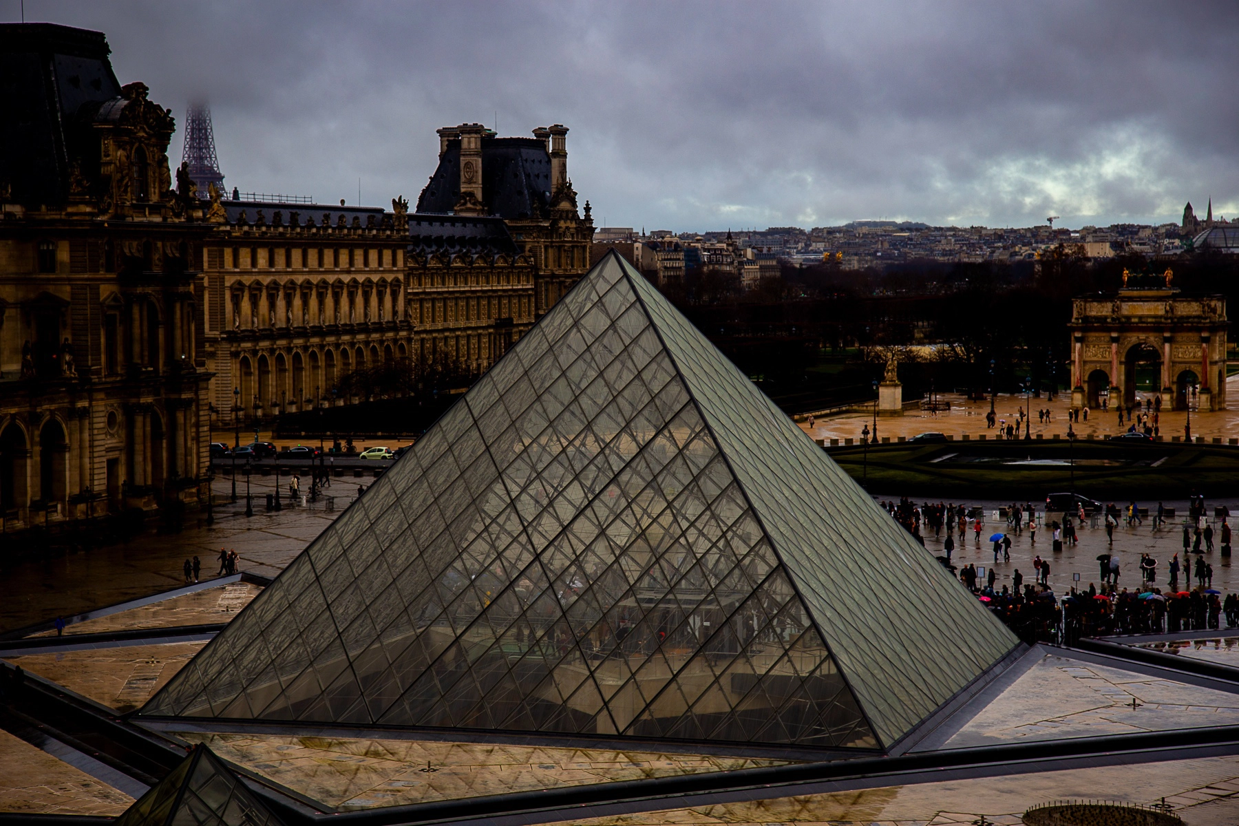 A view of the iconic Louvre Pyramid with the historic Louvre buildings and the Eiffel Tower in the background on a cloudy day. Paris,France